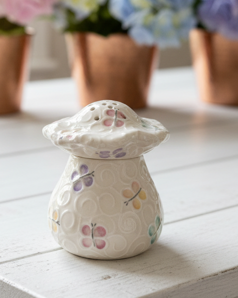 A small white painted butterfly pattern fruit fly trap with a matching lid, decorated with a pastel butterfly and swirl pattern, sitting on a white wooden surface with a blurred background of potted flowers.