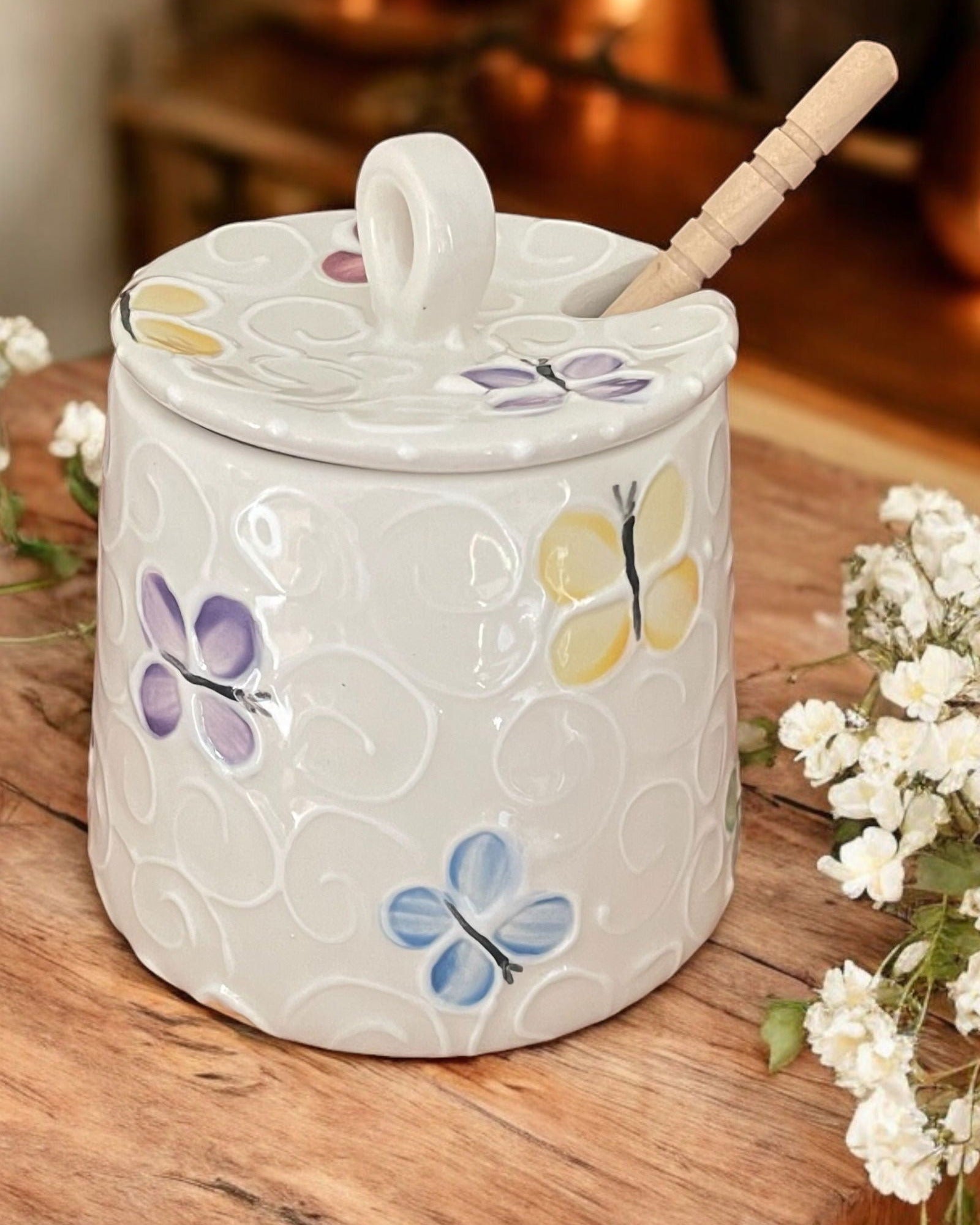 A cream-colored  ceramic honey pot jar with a lid and a wooden spoon, decorated with colorful butterfly motif, set on a rustic wooden table. 