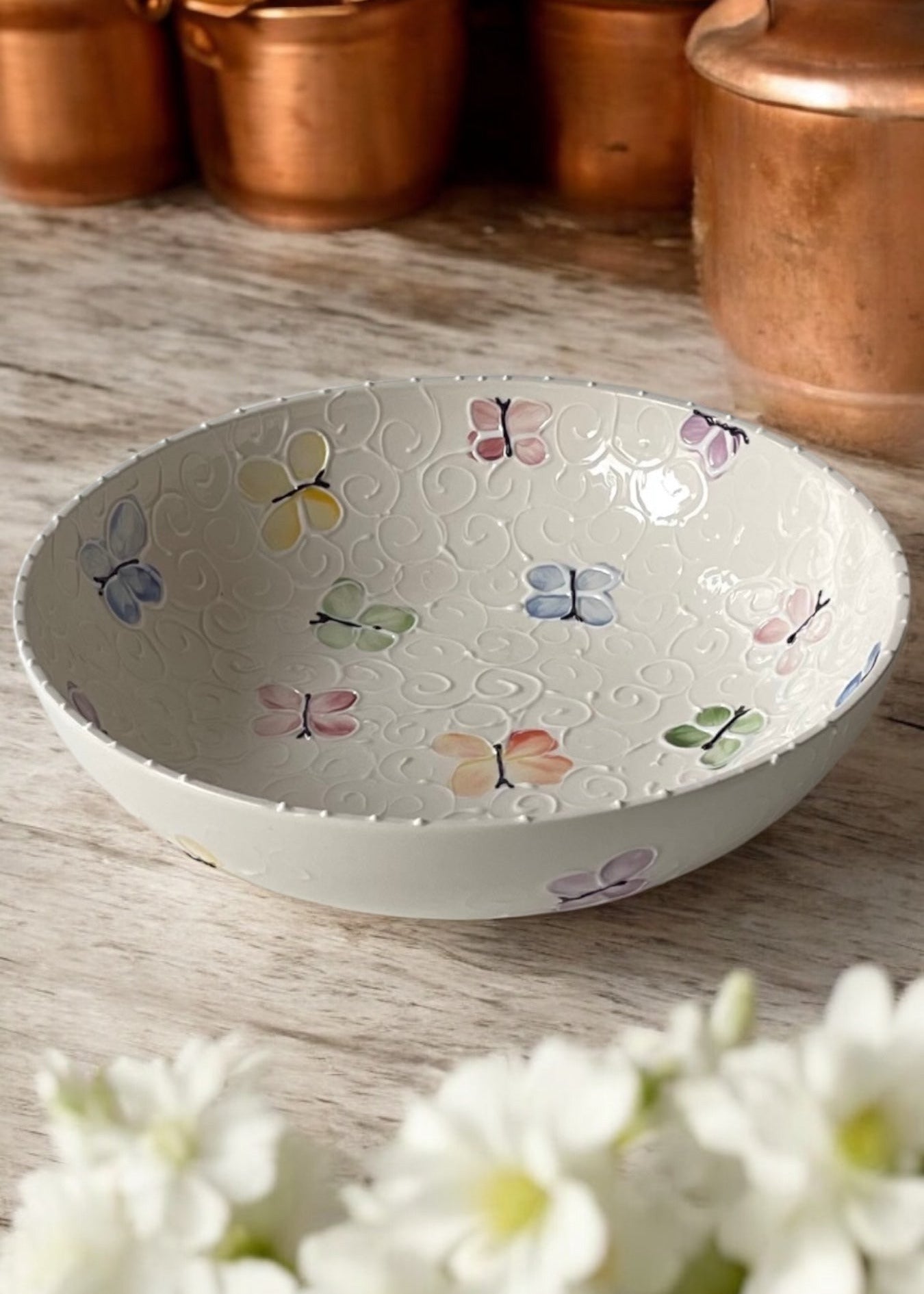 A hand-painted ceramic serving bowl with floral patterns, displayed on a wooden table with daisies in the foreground and copper cookware in the background.