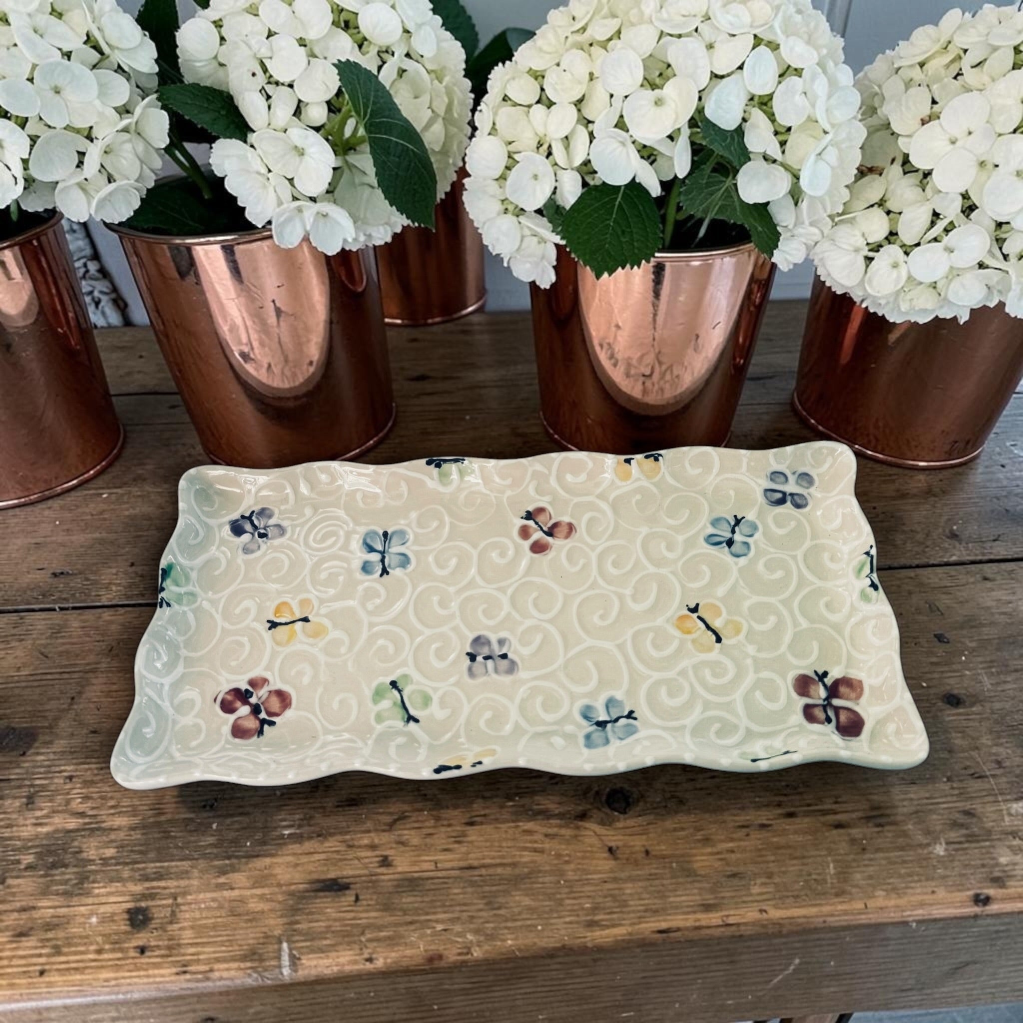 Decorative tray with painted butterfly pattern on a wooden surface with white flowers in copper pots.