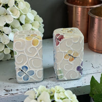 Two decorative ceramic Butterfly salt and pepper shakers with floral patterns on a wooden surface with flowers and copper containers in the background.