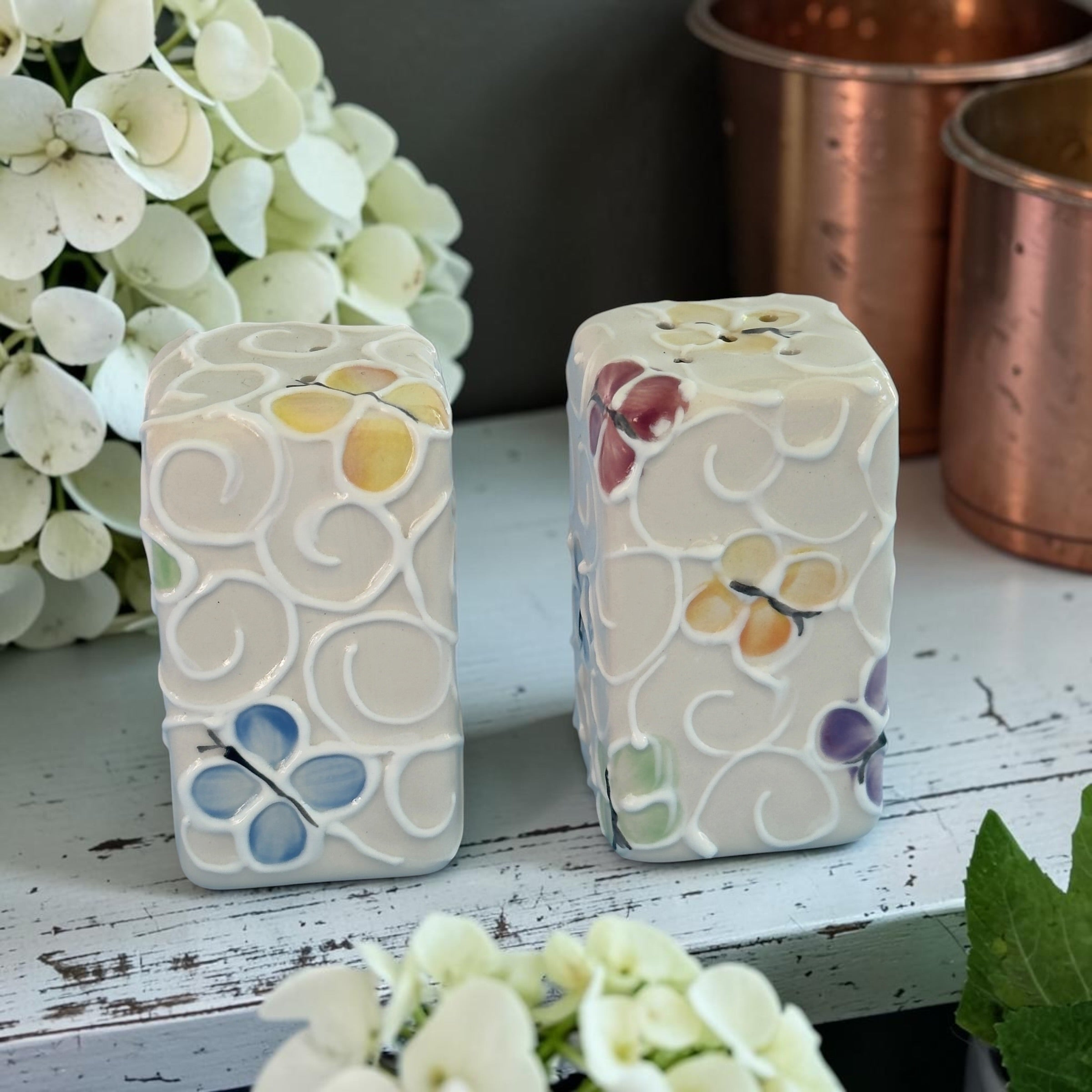 Two decorative ceramic Butterfly salt and pepper shakers with floral patterns on a wooden surface with flowers and copper containers in the background.