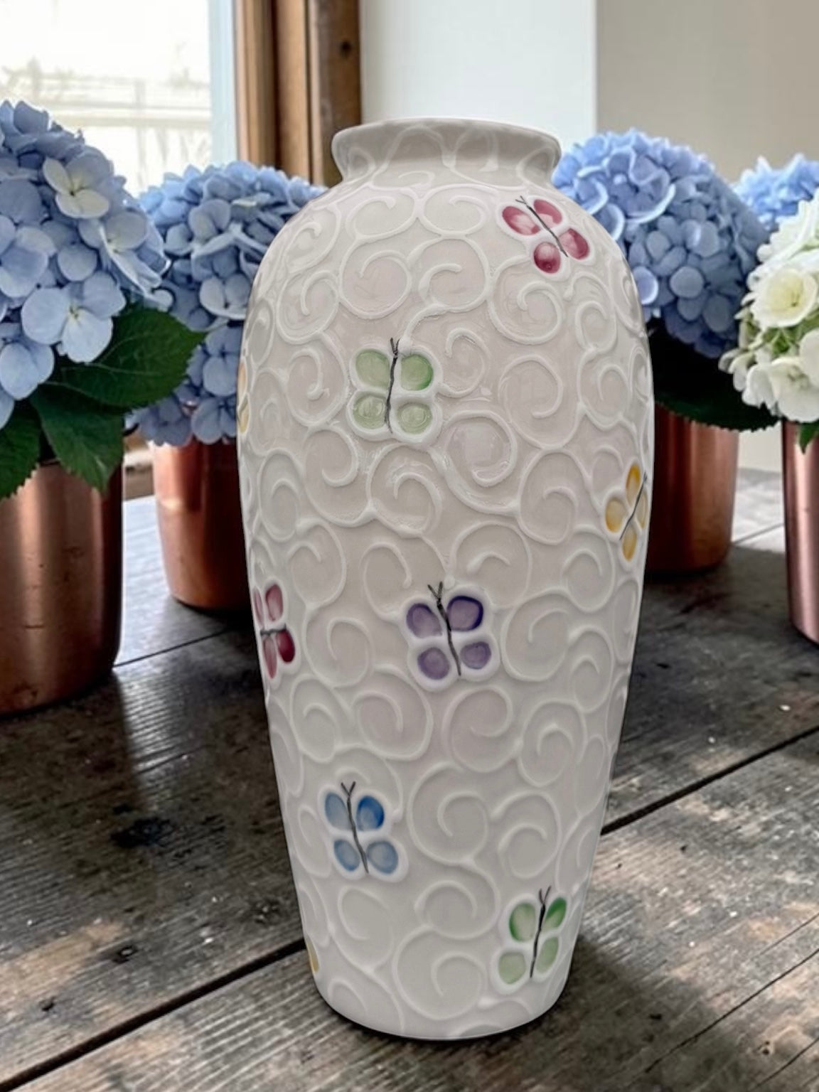 A white ceramic vase with a textured embossed swirl pattern and small hand-painted butterfly motifs in pastel colors, displayed on a rustic wooden table with blue hydrangeas in the background.