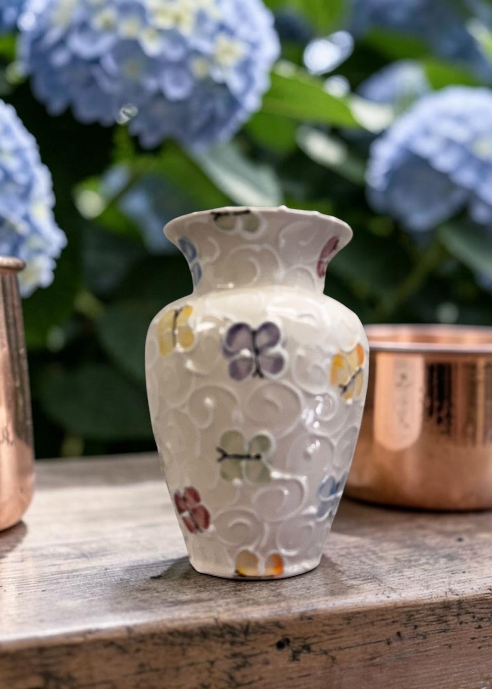 Decorative vase with butterfly painted patterns on a wooden surface with copper pots and blue flowers in the background.