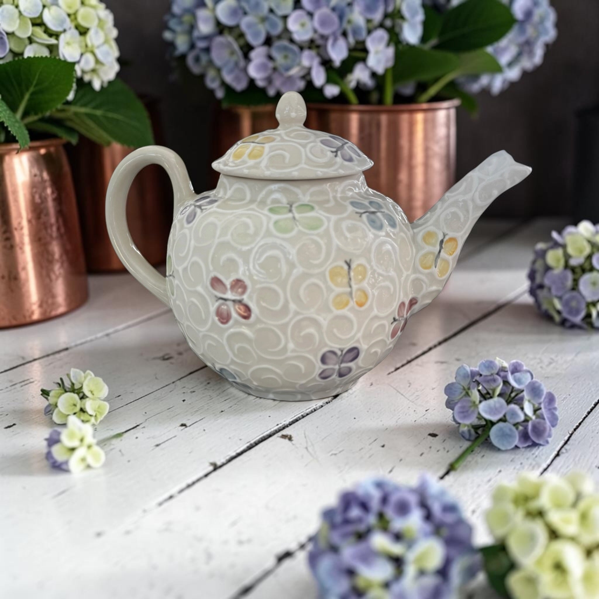 Decorative teapot with butterfly pattern on a wooden surface with flowers around