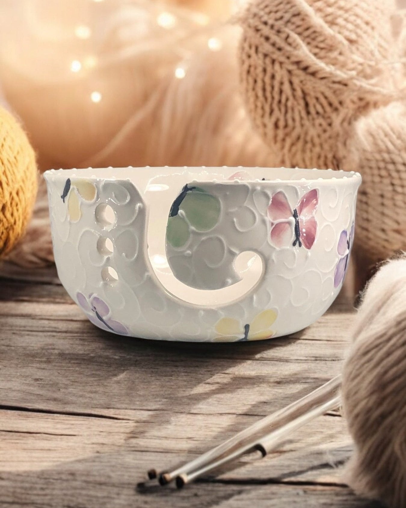 A hand-painted ceramic knitting bowl with butterfly designs, containing yarn balls and knitting tools, placed on a wooden surface. Ladybird Ceramics Canada