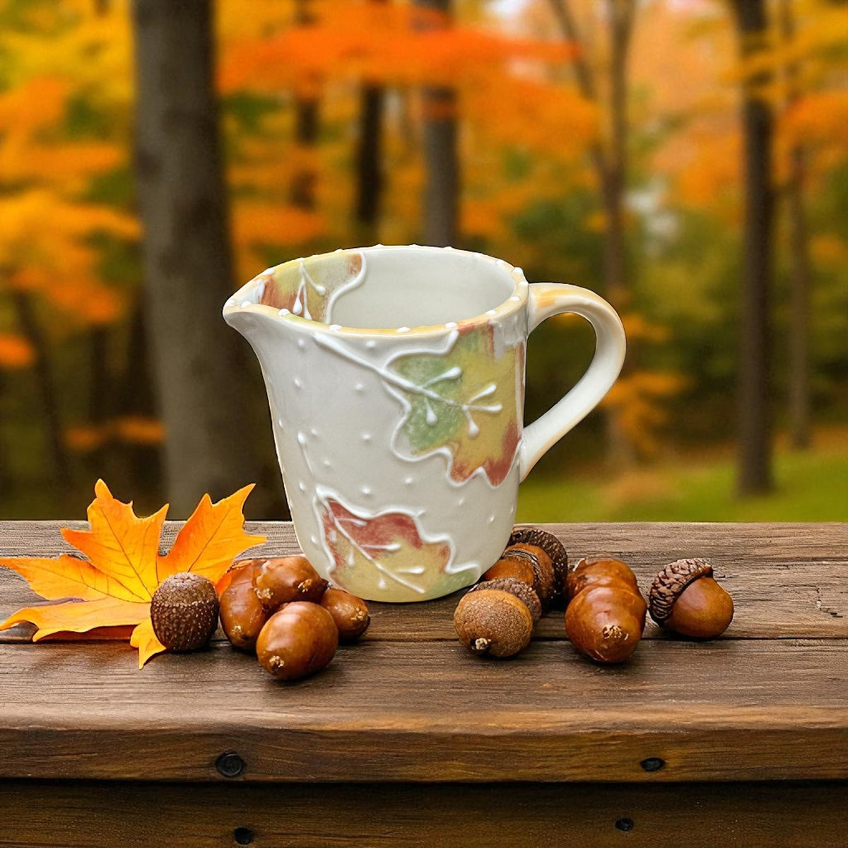 Ceramic milk creamer with handle with autumn leaf design on a wooden surface with acorns and leaves, set against an autumn forest background.