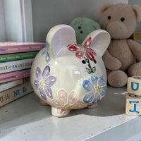 Decorative daisy painted piggy bank with floral designs on a shelf with books and a teddy bear in the background - Ladybird Ceramics