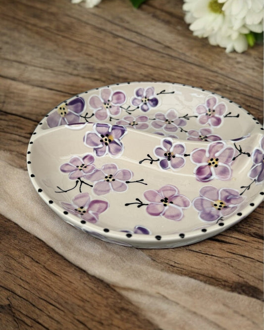 A ceramic two-compartment serving Bowl with a white base decorated with a colorful  purple floral pattern, set on a white wooden surface with potted flowers in the background.