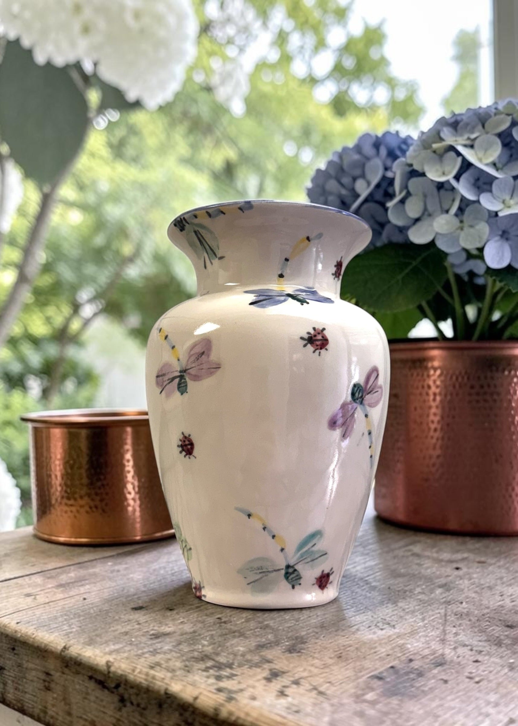 Decorative vase with dragonfly patterns on a wooden surface with hydrangeas in the background