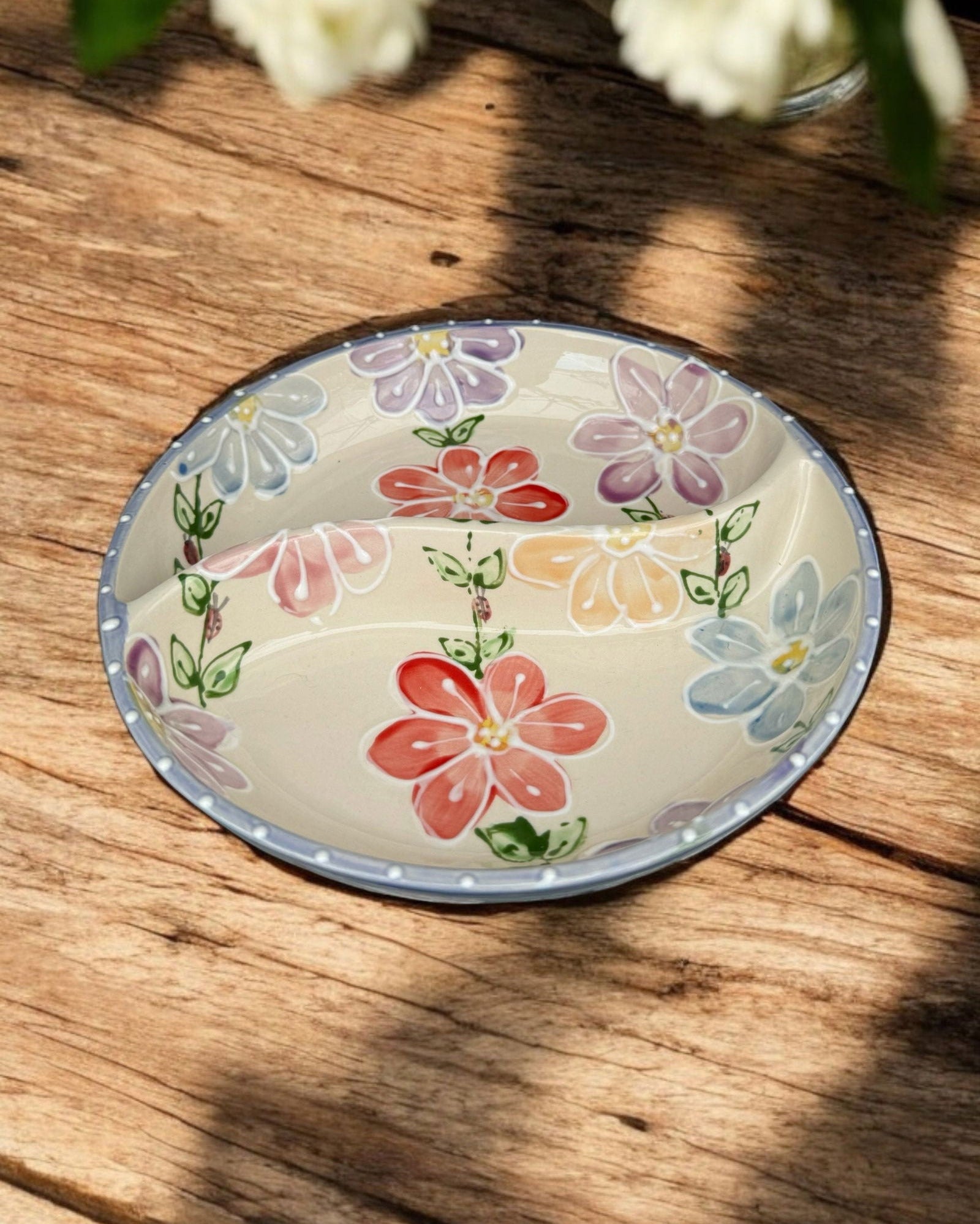 A ceramic two-compartment serving Bowl with a white base decorated with a colorful floral pattern, set on a white wooden surface with potted flowers in the background.