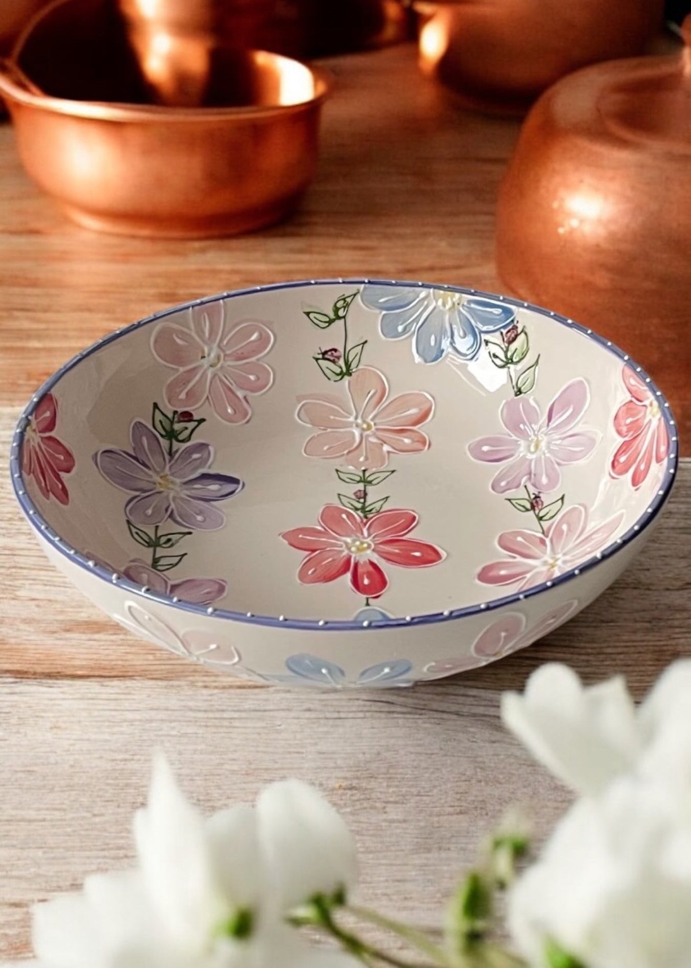 A hand-painted ceramic serving bowl with floral patterns, displayed on a wooden table with daisies in the foreground and copper cookware in the background.
