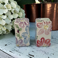 Two floral-patterned ceramic salt and pepper blocks on a wooden surface with white flowers and a copper pot in the background.