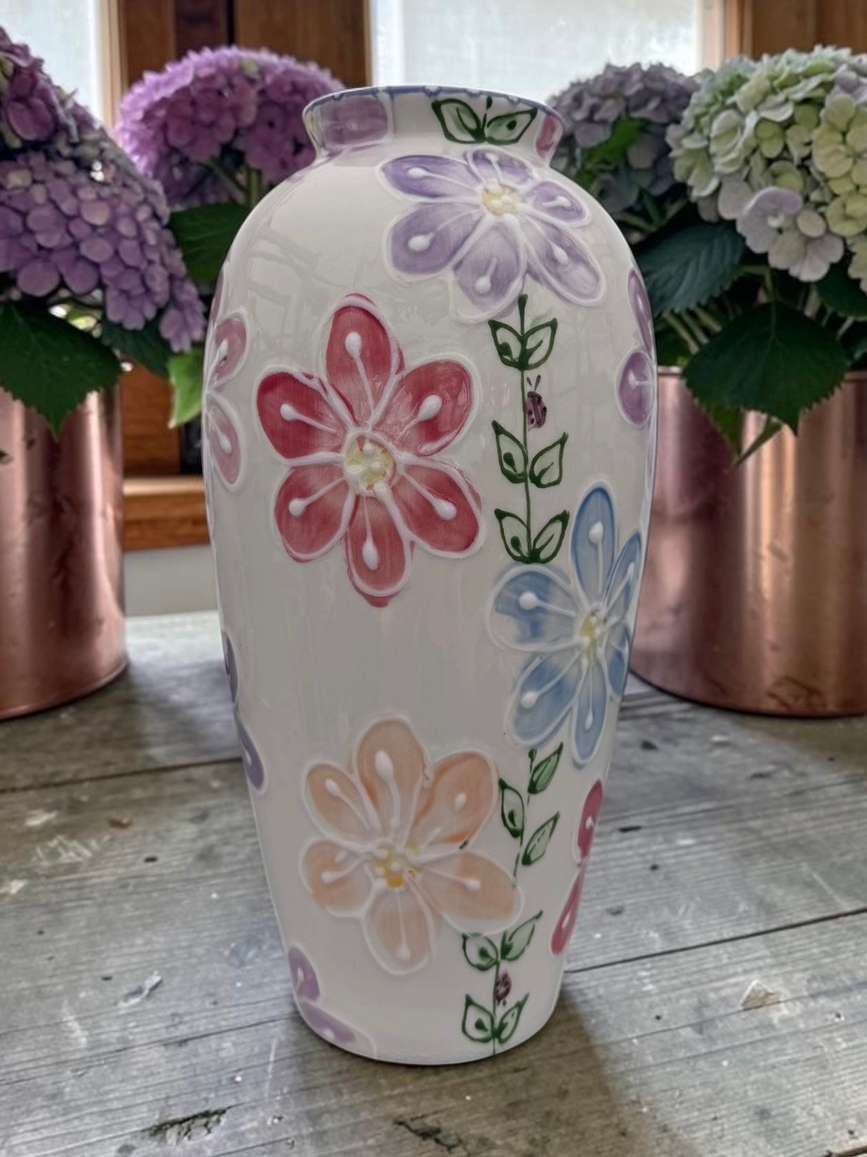A white ceramic vase decorated with hand-painted pastel flowers in pink, purple, blue, and peach, displayed on a rustic wooden table with copper planters and hydrangeas in the background.