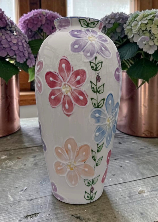 A white ceramic vase decorated with hand-painted pastel flowers in pink, purple, blue, and peach, displayed on a rustic wooden table with copper planters and hydrangeas in the background.
