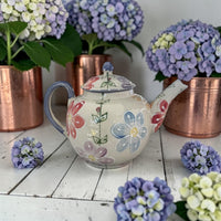 Floral teapot daisy pattern on a wooden surface with potted hydrangeas in the background