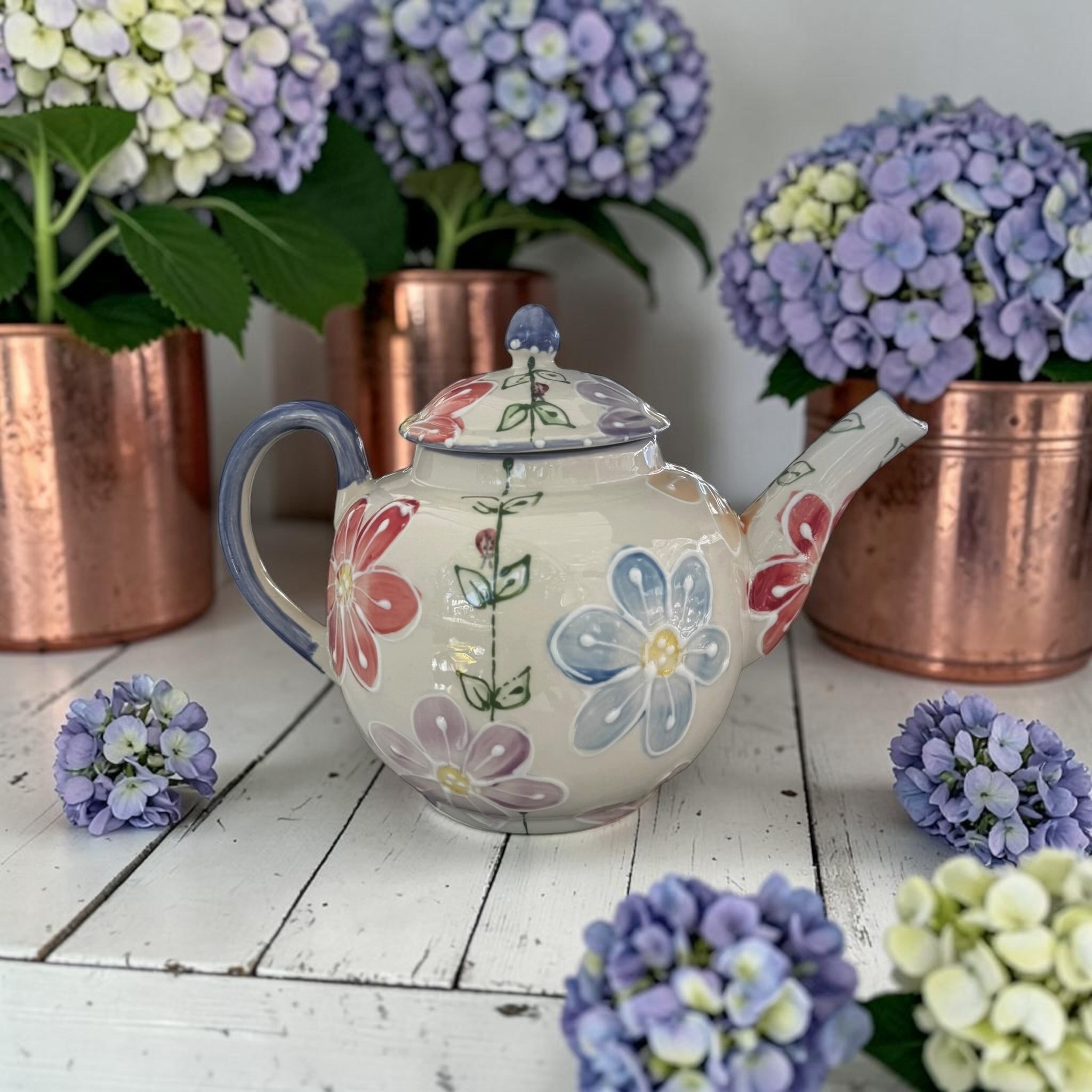 Floral teapot daisy pattern on a wooden surface with potted hydrangeas in the background