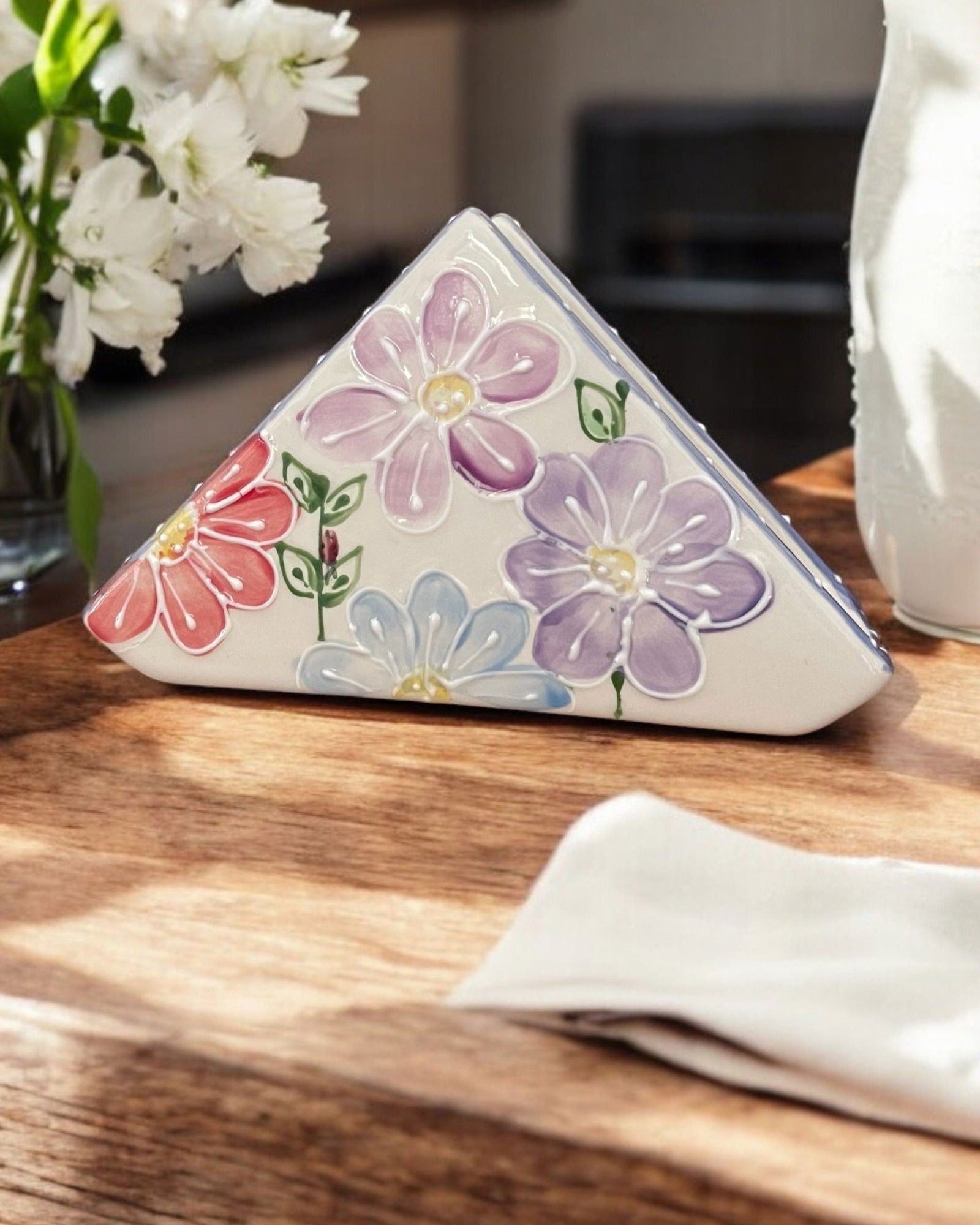 A triangular white ceramic napkin holder with painted  floral patterns displayed on a wooden table.