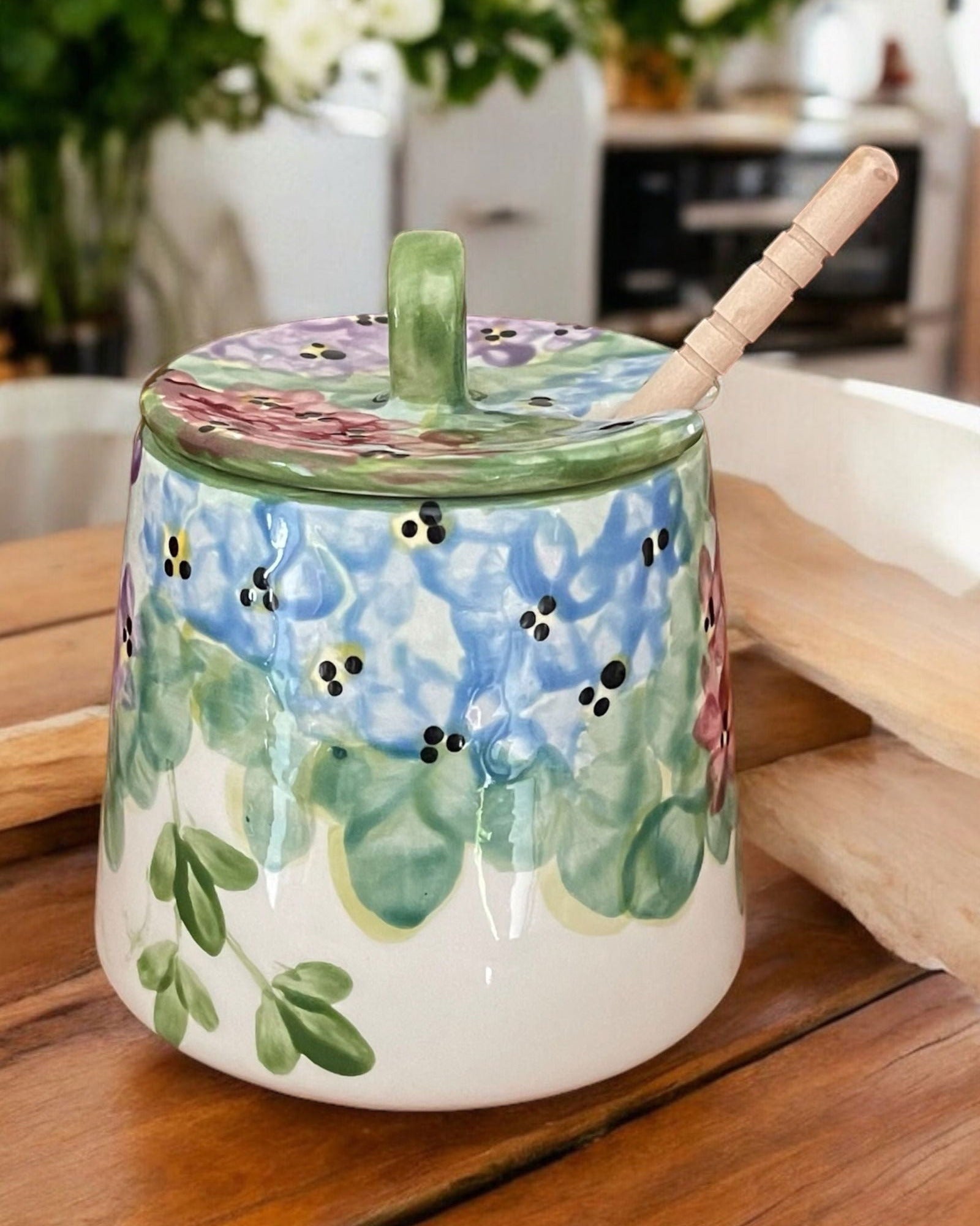 A ceramic honey pot with a lid, decorated with blue flowers, green leaves, and black and white details, sitting on a wooden table with a wooden spoon inside.