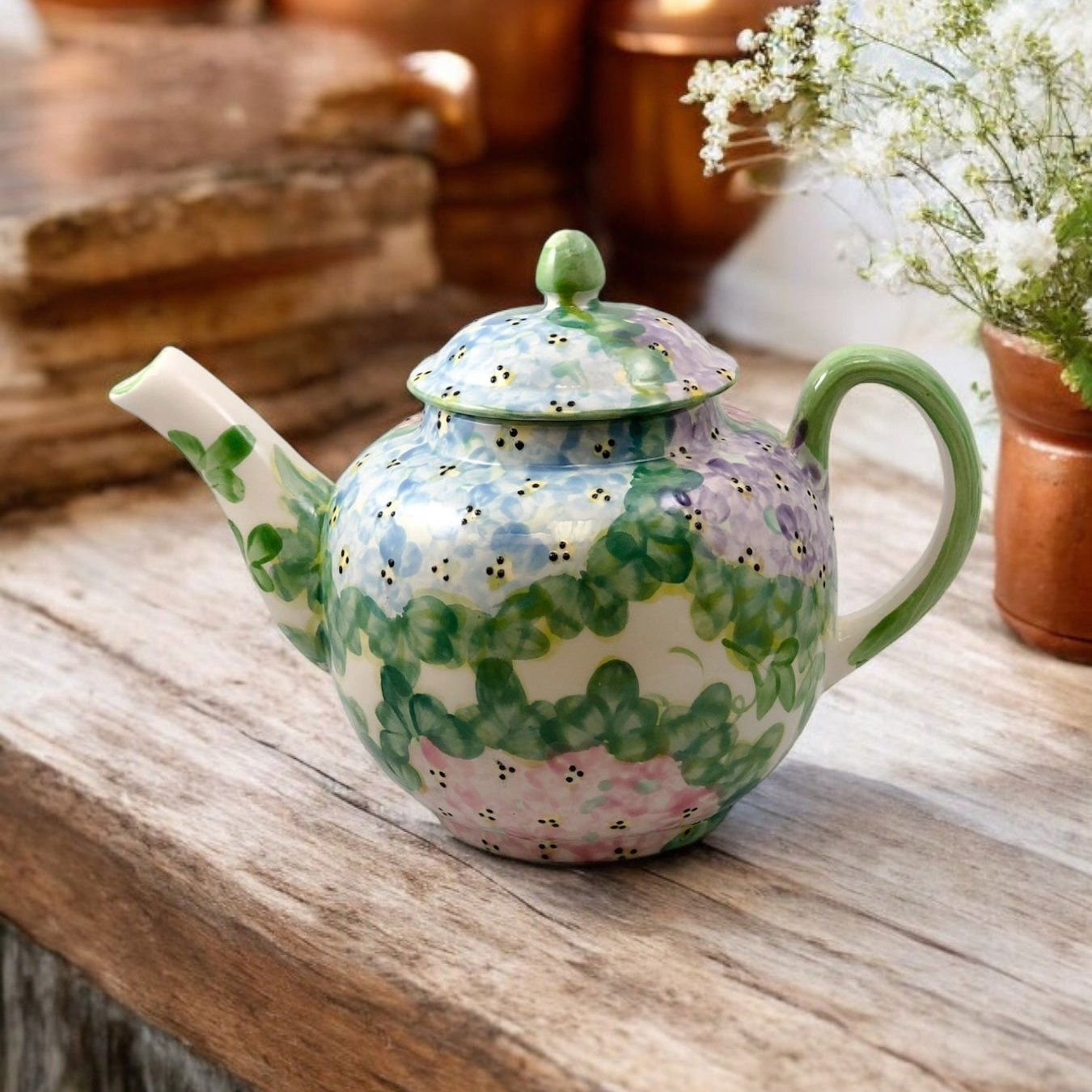 A white ceramic teapot with hand-painted floral designs, displayed on a wooden surface with a bouquet of white flowers beside it.