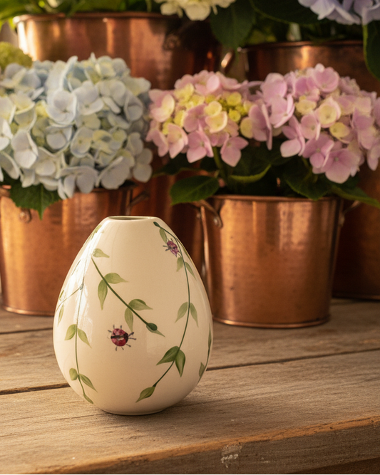 A small cream-colored ceramic vase with a painted green vine and ladybug design sits on a rustic wooden table, with copper planters and pastel hydrangeas blurred in the background.