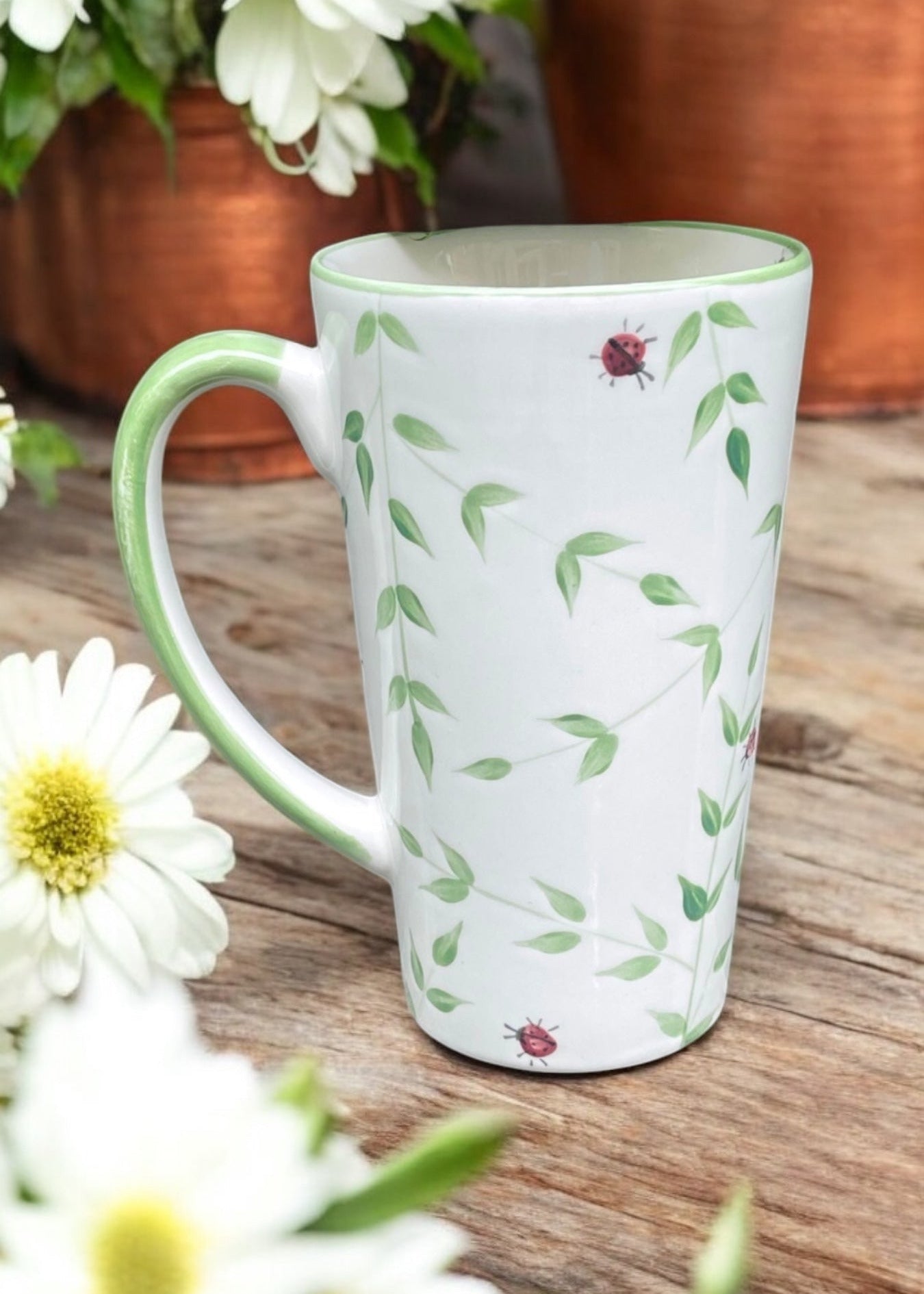 A white ceramic mug with a green handle and rim, decorated with a green leaf vine pattern and small ladybugs. The mug is set on a rustic wooden surface with white daisies and copper pots blurred in the background.
