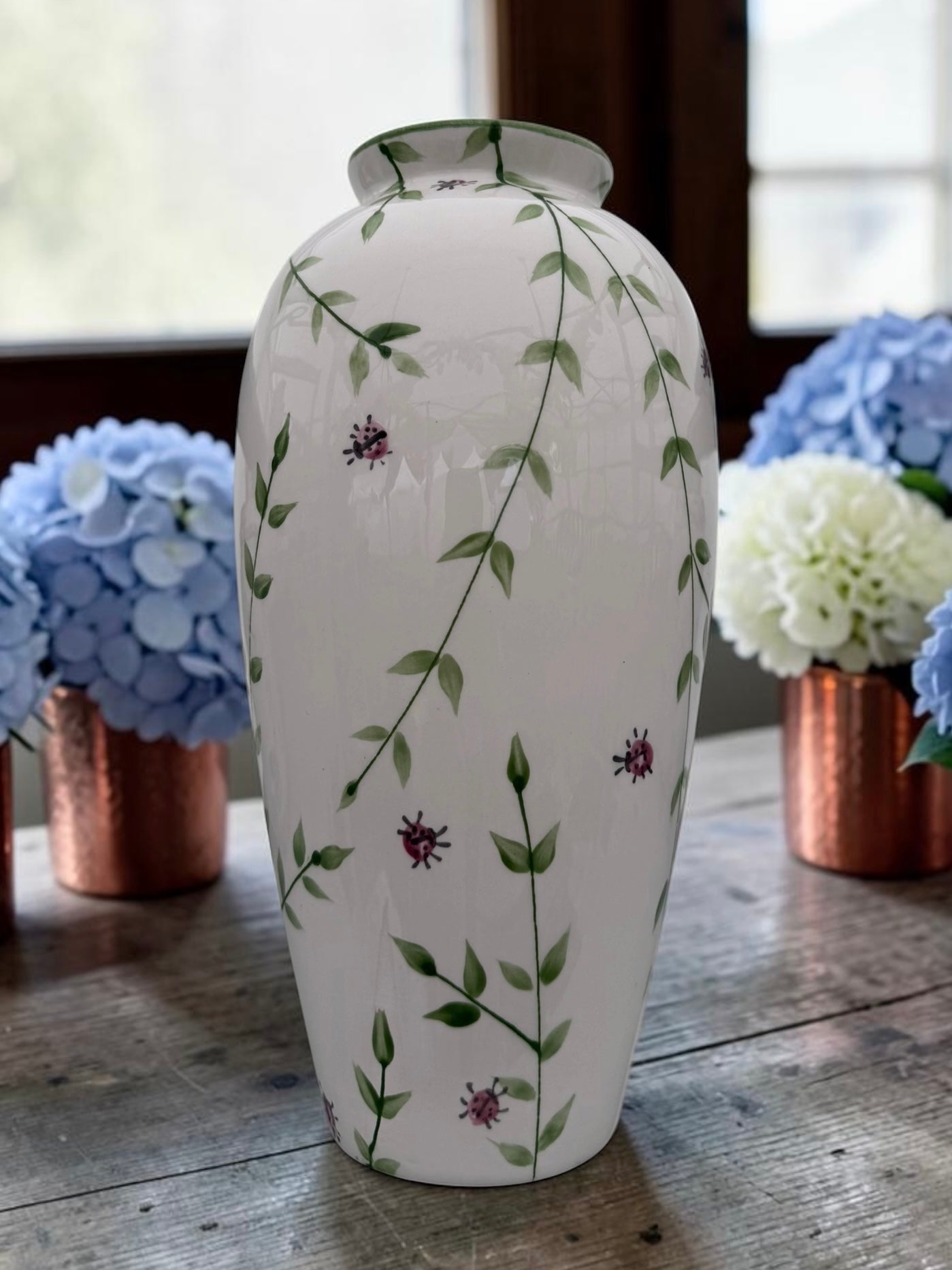A white ceramic vase decorated with green leafy vines and small purple flowers, displayed on a rustic wooden table with blue and white hydrangeas in the background.