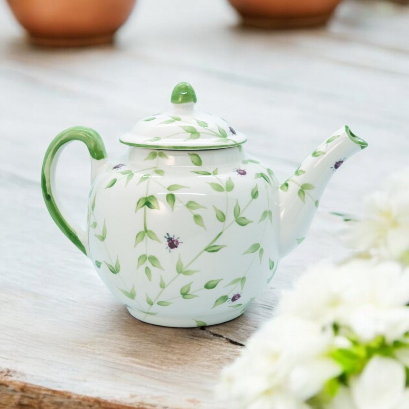 A white ceramic teapot with hand-painted floral designs, displayed on a wooden surface with a bouquet of white flowers beside it.