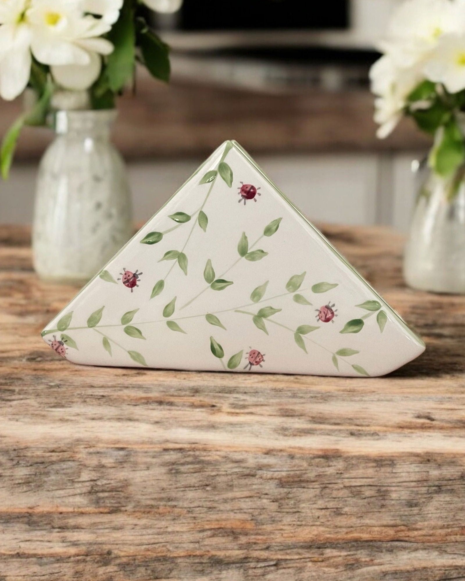 A triangular white ceramic napkin holder with painted green leaf floral patterns displayed on a wooden table.