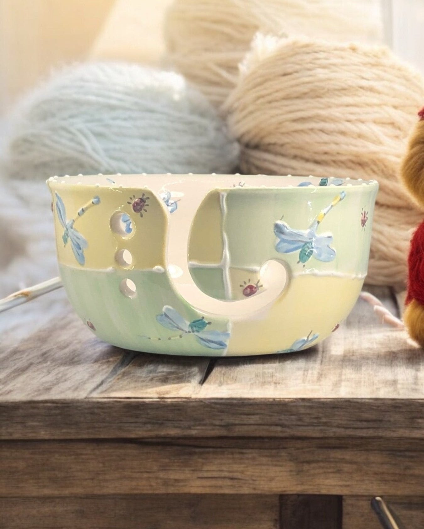 A hand-painted ceramic knitting bowl with yellow dragonfly designs, containing yarn balls and knitting tools, placed on a wooden surface. Ladybird Ceramics Canada
