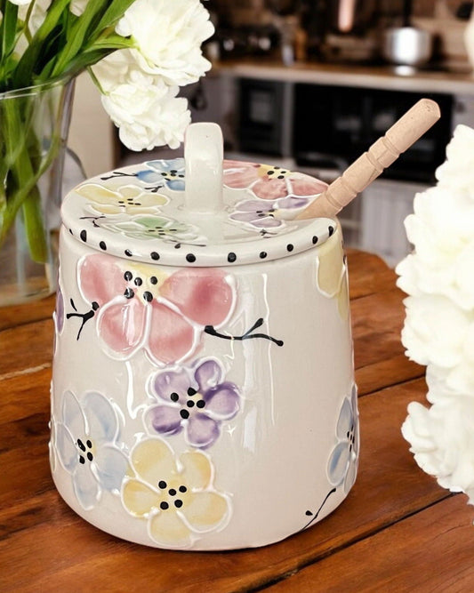 A white ceramic honey pot with colorful cherry blossom designs, a wooden spoon, and white flowers on a wooden table.