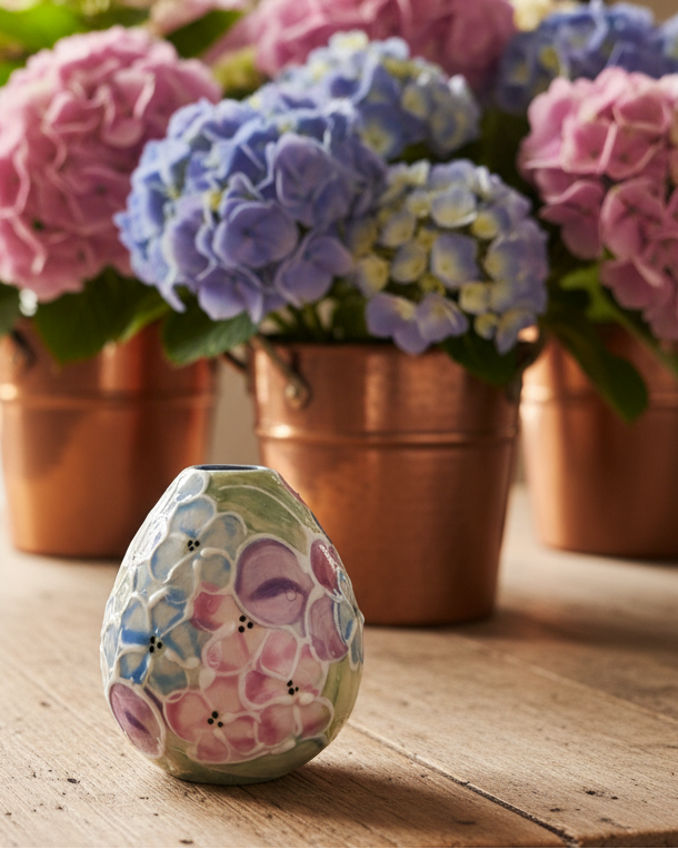 A small decorative ceramic vase with a floral pattern in pastel pink, purple, blue, and green, placed on a wooden table in front of potted hydrangeas in copper pots.