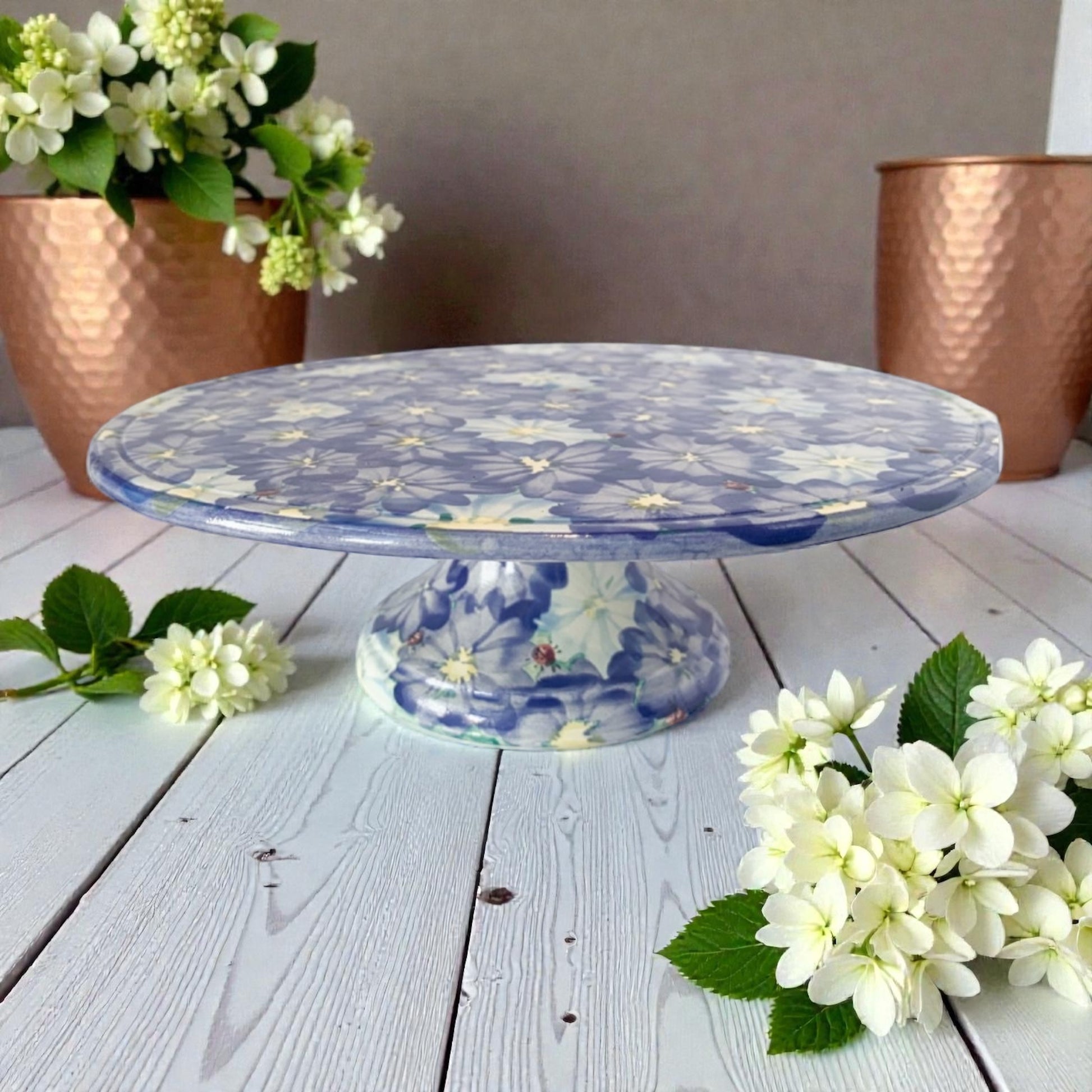 Decorative cake stand with blue hydrangea floral pattern on a wooden surface with flowers.