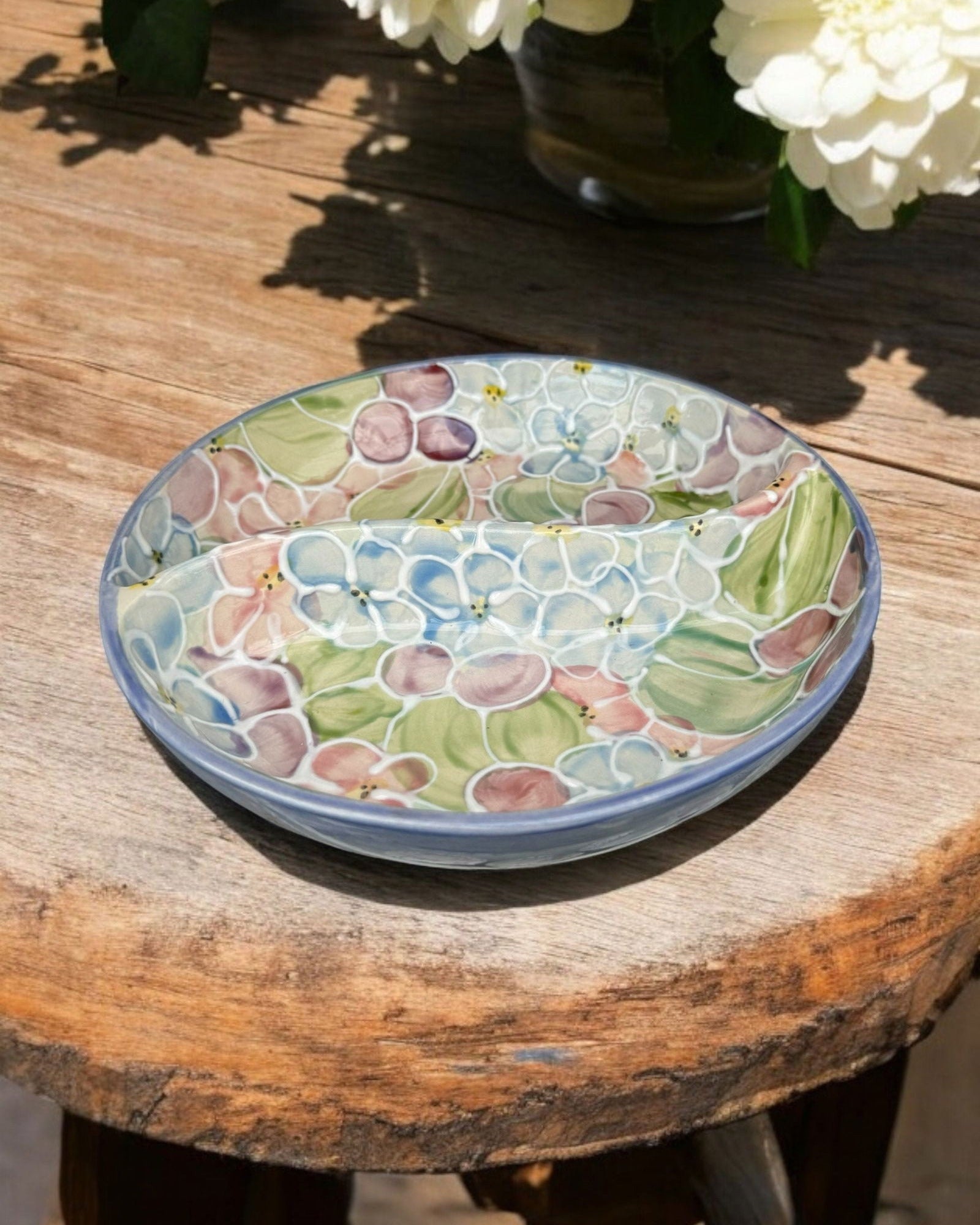 A ceramic two-compartment serving Bowl with a white base decorated with a hydrangea floral pattern, set on a white wooden surface with potted flowers in the background.