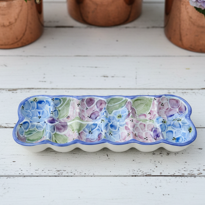 A rectangular ceramic dish with a wavy scalloped rim, decorated with a floral pattern in shades of blue, purple, and green on a white base. It is styled on a white wooden surface with copper pots of purple hydrangeas in the background.