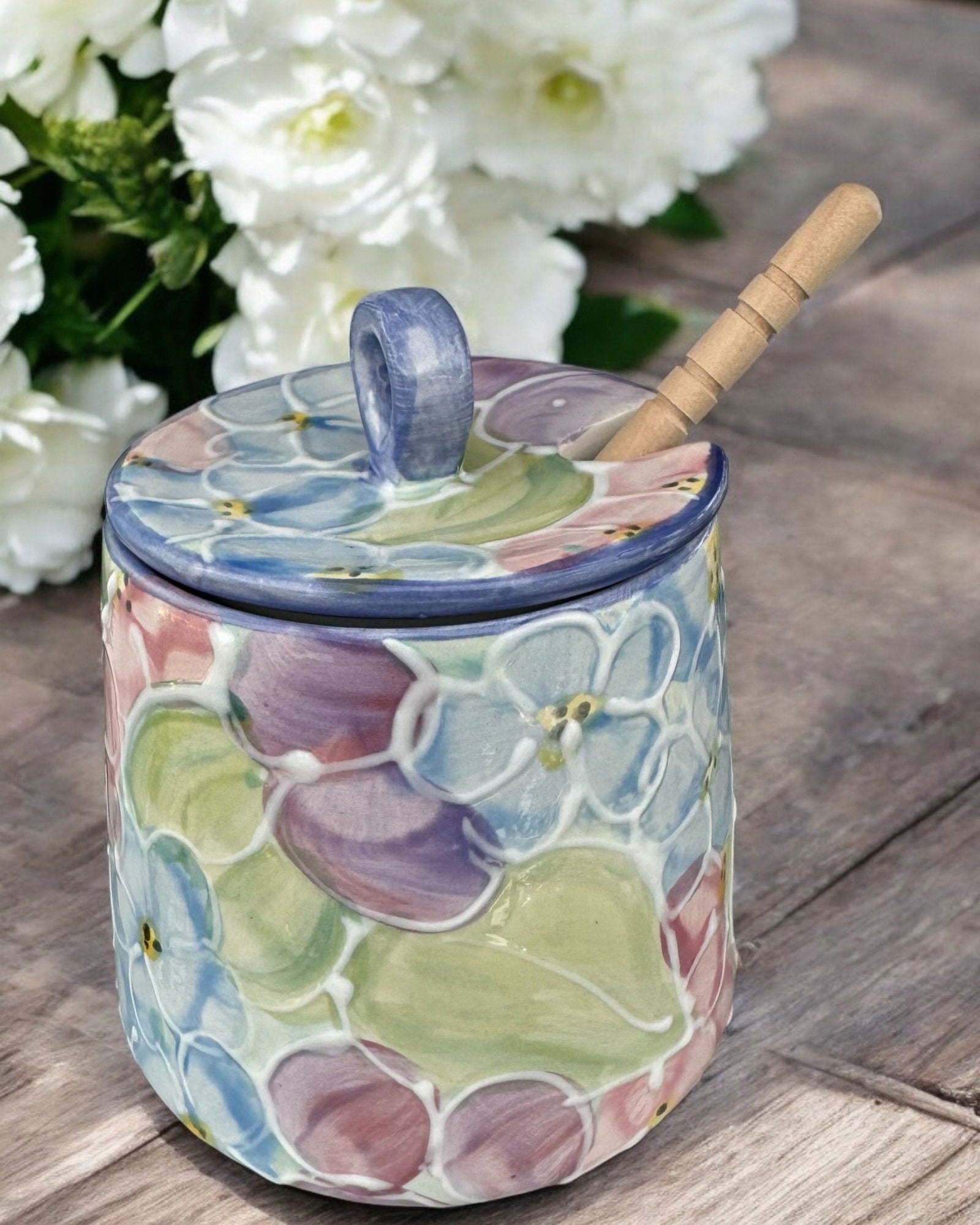 A cream-colored  ceramic honey pot jar with a lid and a wooden spoon, decorated with colorful hydrangea motifs, set on a rustic wooden table with blurred purple and blue hydrangeas in the background.