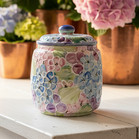 A small ceramic jar with a matching lid, decorated with a hand-painted floral pattern in pastel blue, pink, purple, and green, sitting on a white wooden surface with blurred hydrangea bouquets in the background.