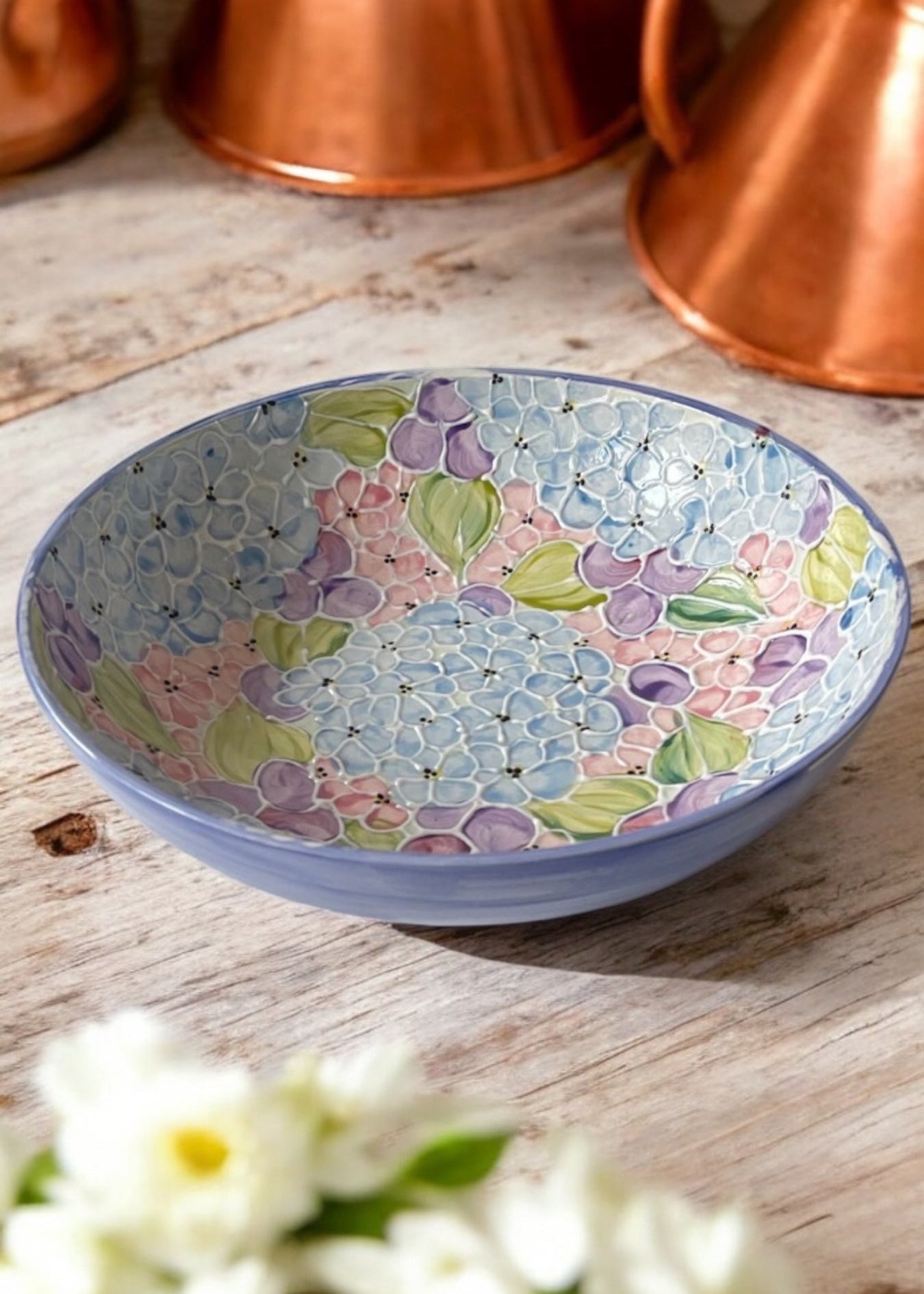 A hand-painted ceramic serving bowl with floral patterns, displayed on a wooden table with daisies in the foreground and copper cookware in the background.