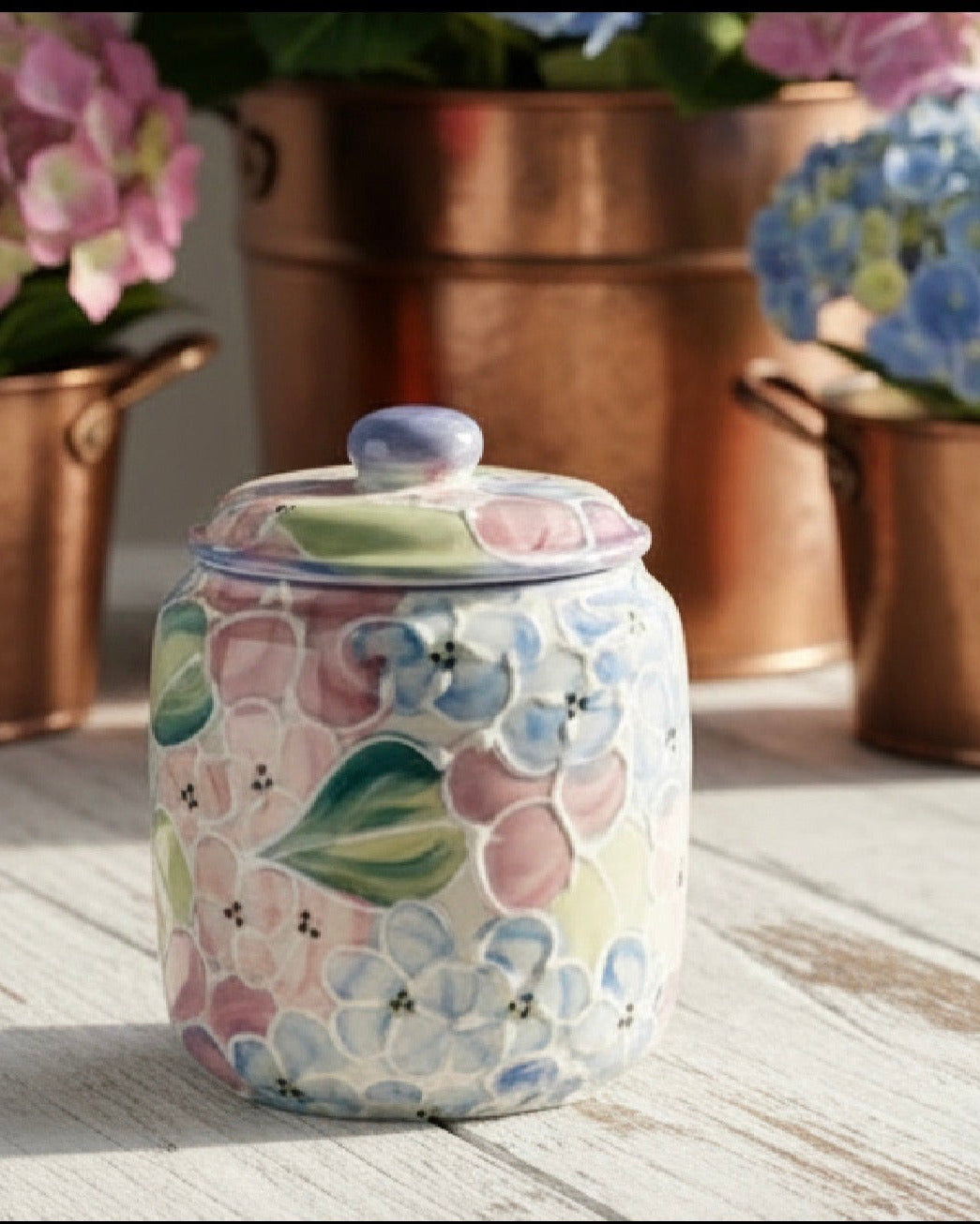 A ceramic lidded jar with a floral pattern in pastel pink, blue, green, and cream, set on a white wooden surface with blurred copper planters and flowers in the background.