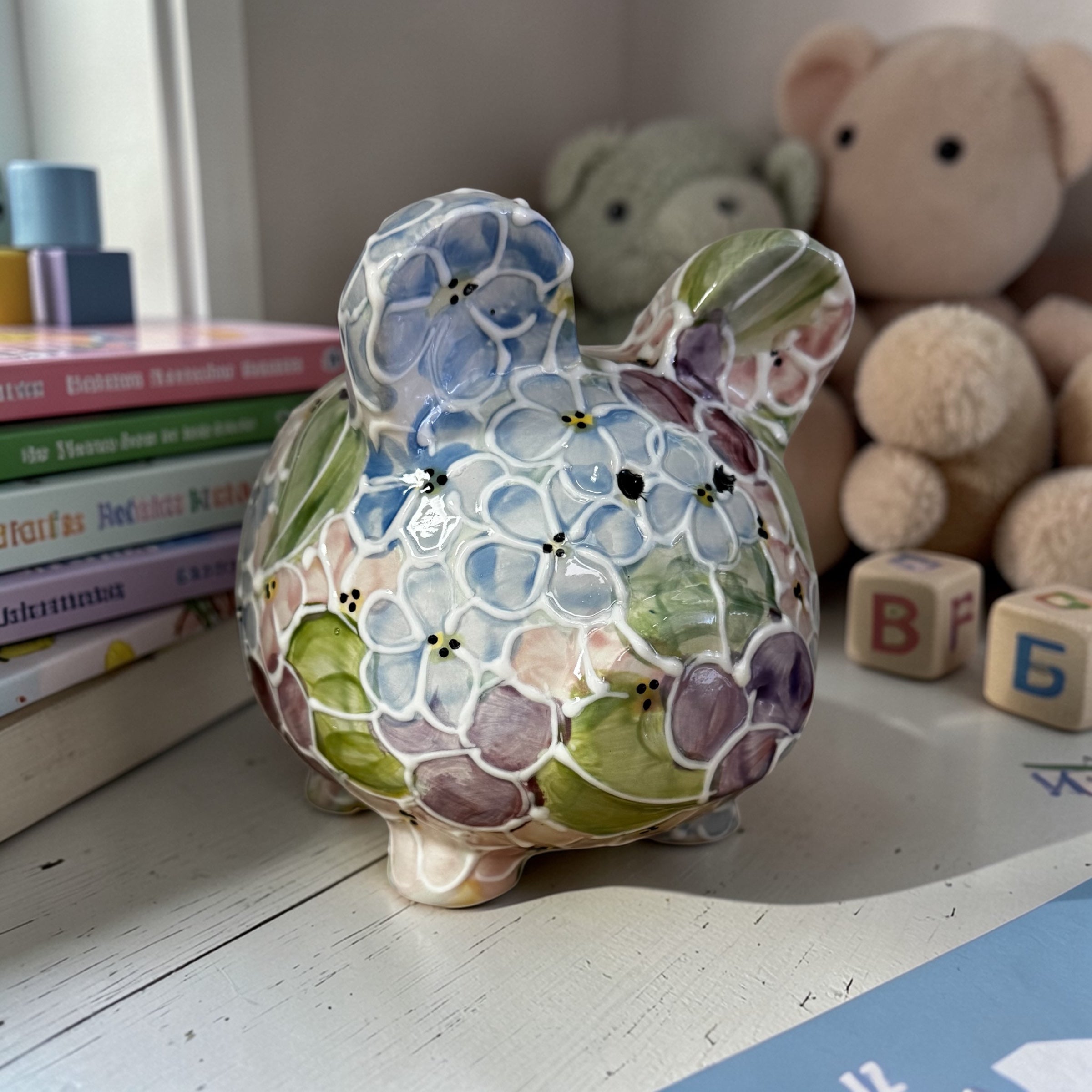 Floral-patterned hydrangea piggy bank on a surface with books, toys, and blocks in the background - Ladybird Ceramics