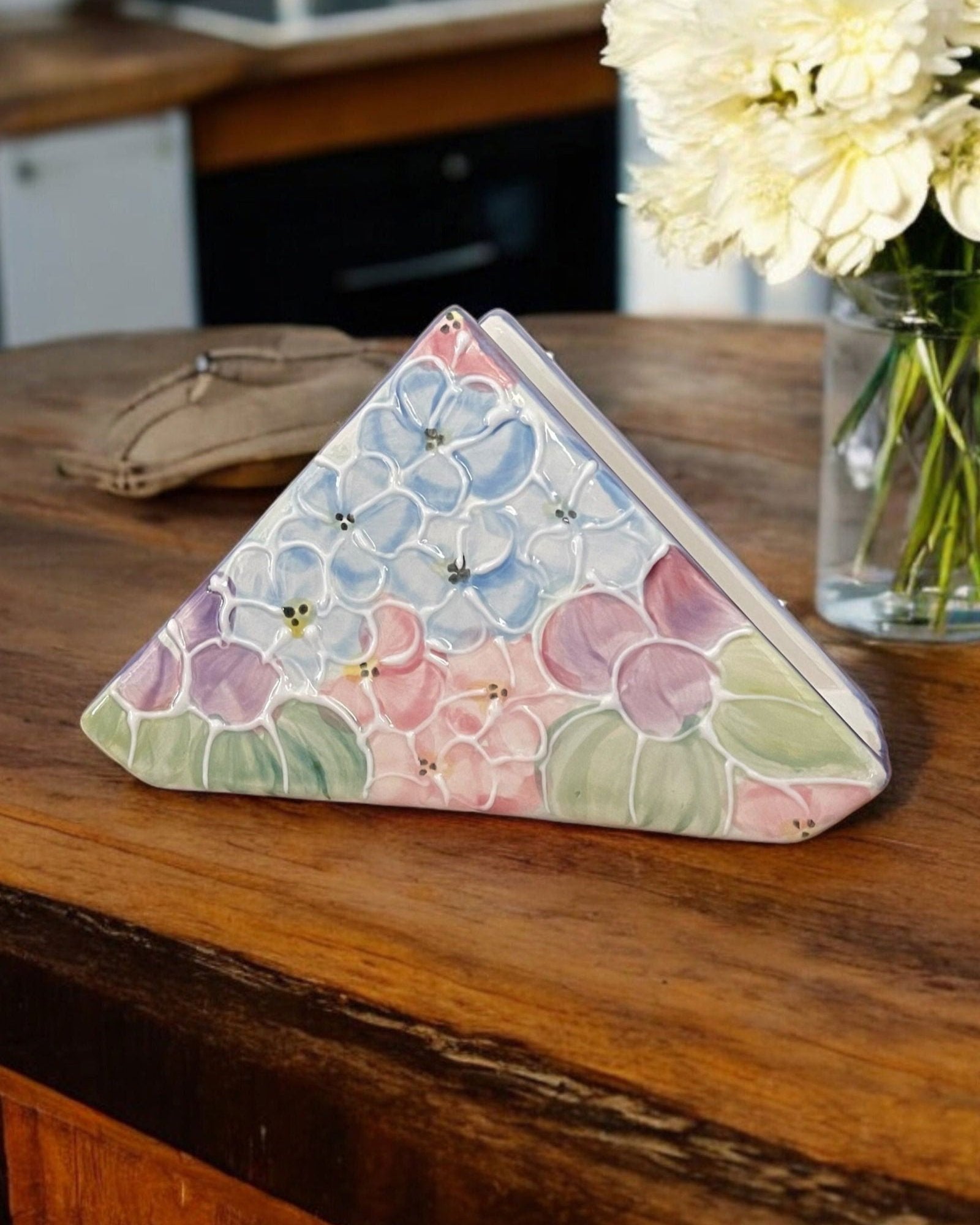 A triangular white ceramic napkin holder with embossed hydrangea floral patterns displayed on a wooden table.