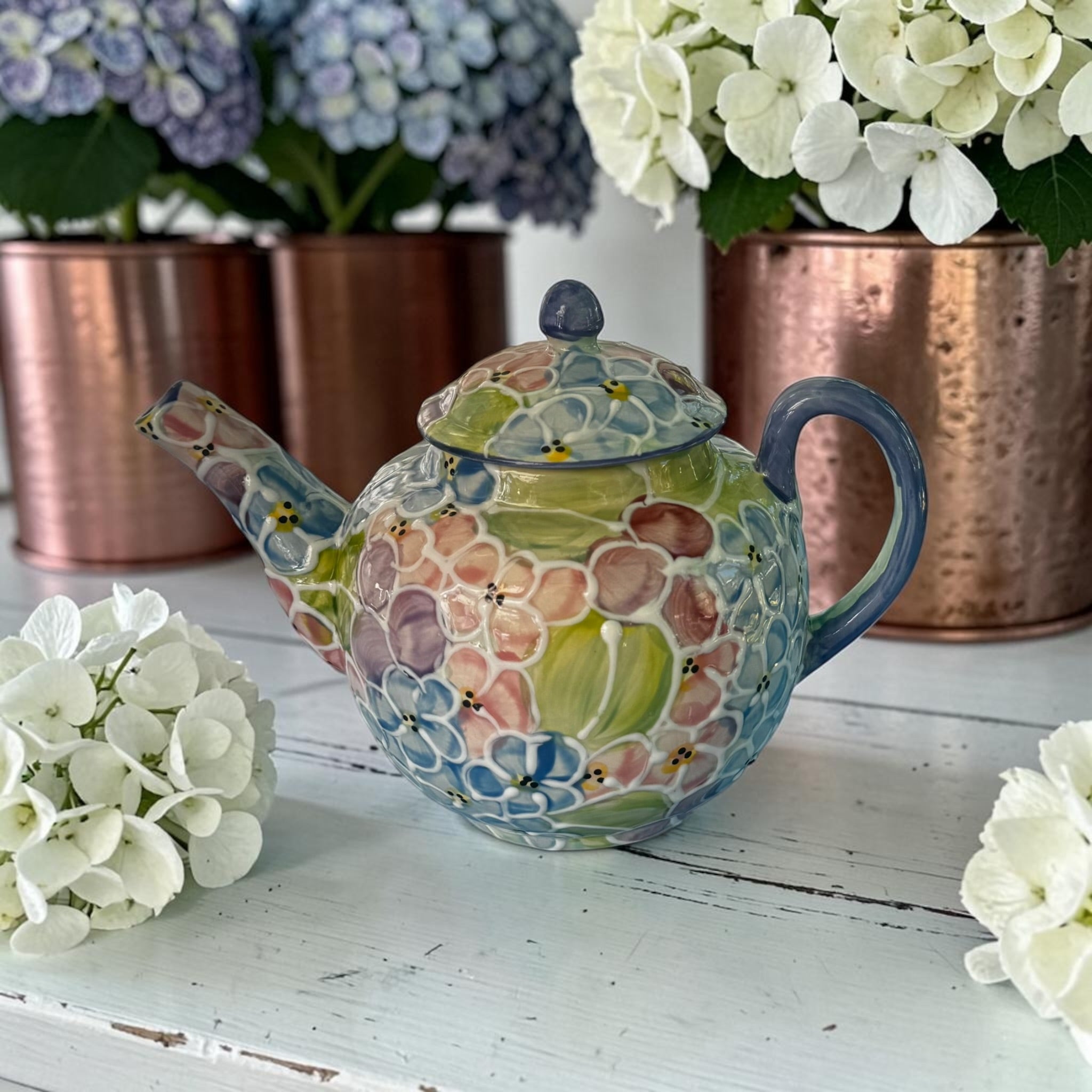 Decorative teapot with floral pattern on a white surface with flowers and copper pots in the background