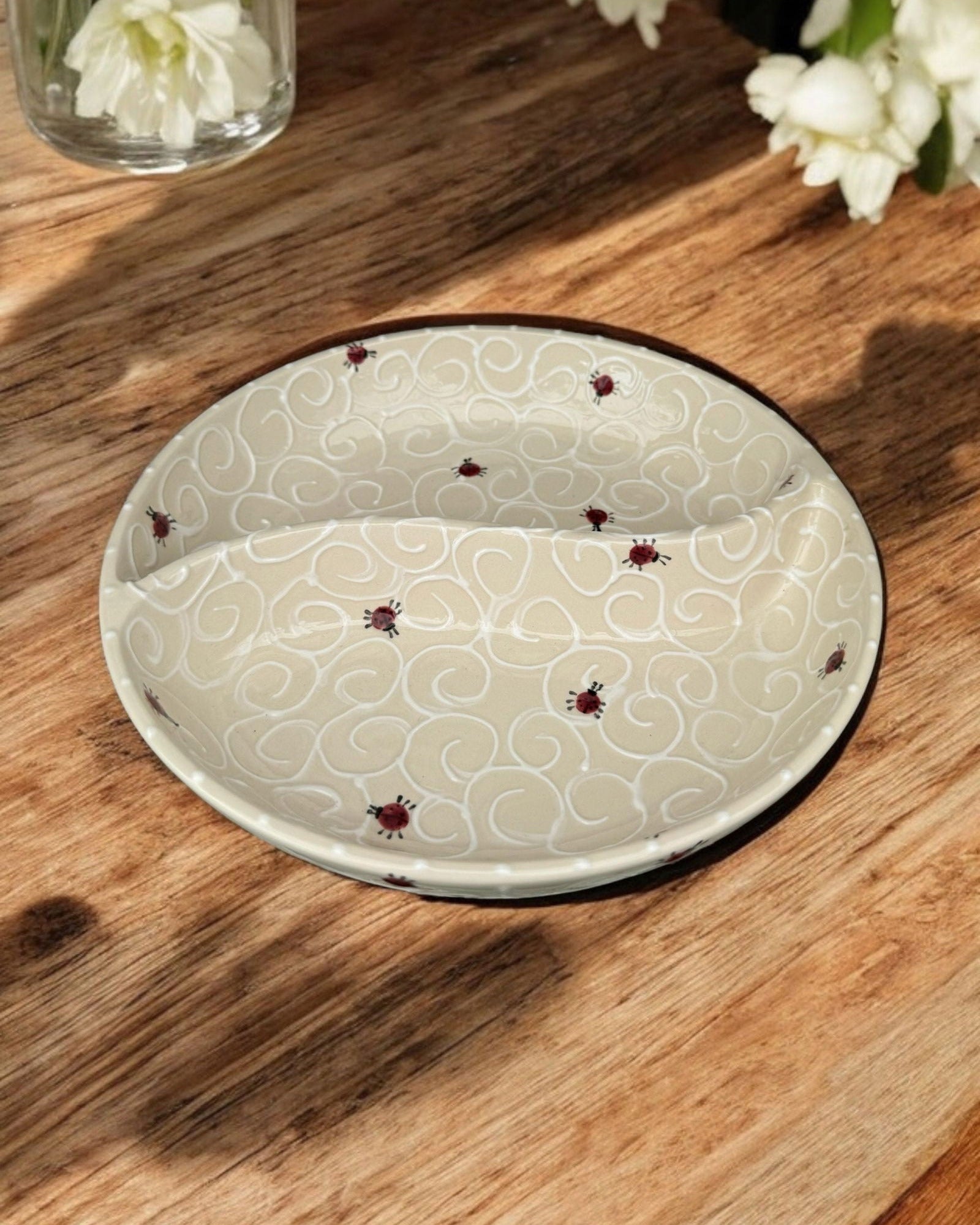 A ceramic two-compartment serving Bowl with a white base decorated with a colorful ladybug pattern, set on a white wooden surface with potted flowers in the background.