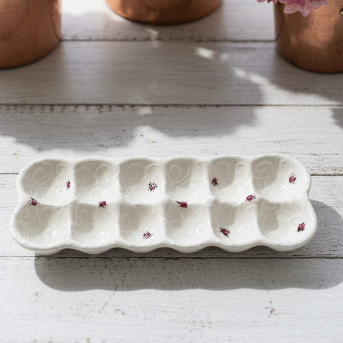 A white ceramic egg-shaped tray with small pink ladybugs on a white wooden table, with copper pots of pink and white hydrangeas in the background.