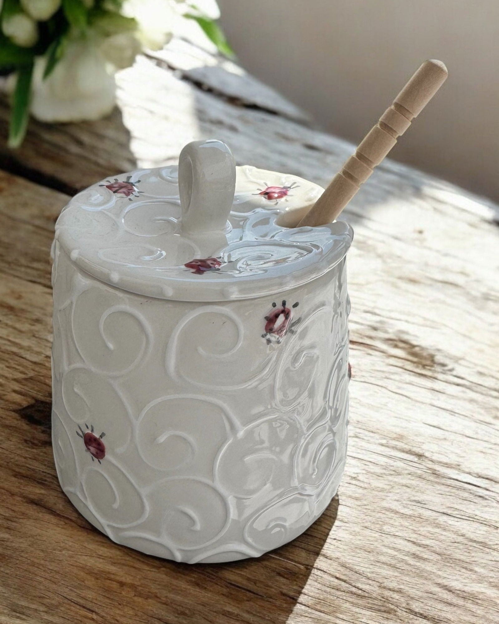 A cream-colored  ceramic honey pot jar with a lid and a wooden spoon, decorated with colorful wildflower and insect motifs, set on a rustic wooden table with blurred purple and blue hydrangeas in the background.