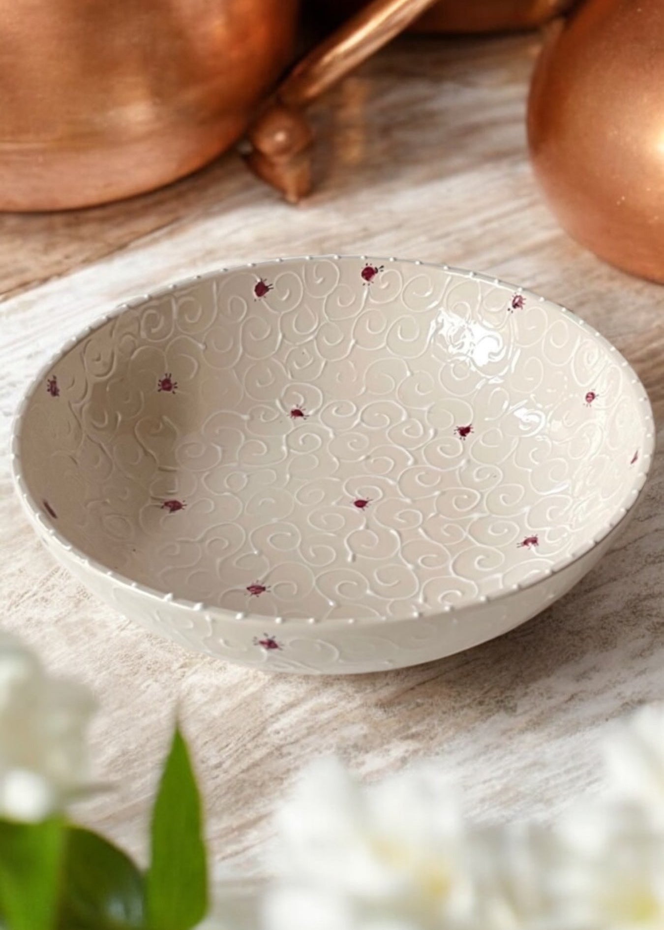 A hand-painted ceramic serving bowl with floral patterns, displayed on a wooden table with daisies in the foreground and copper cookware in the background.
