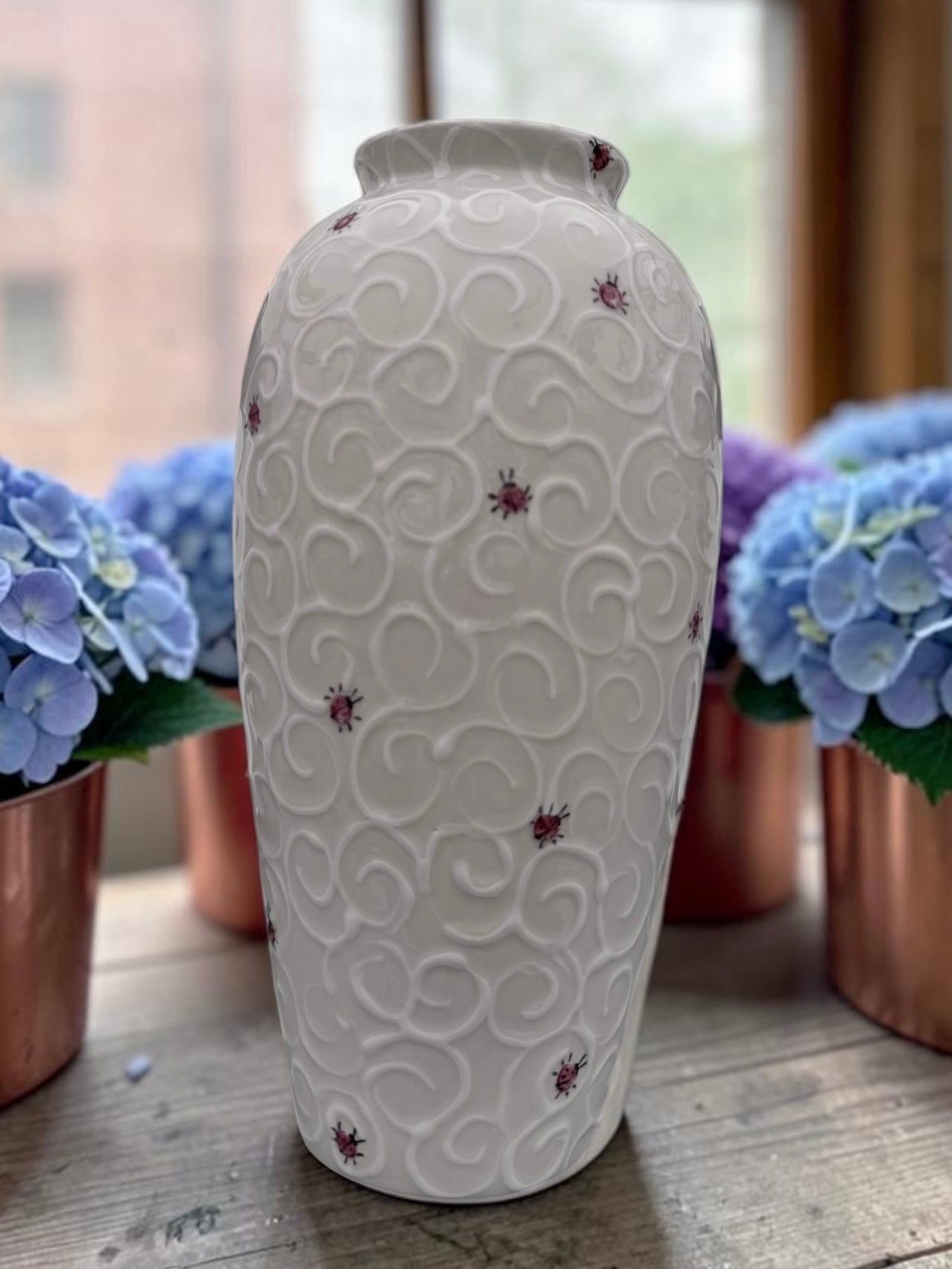 A white ceramic vase with a textured embossed swirl pattern and small red star accents, displayed on a wooden surface with blue hydrangeas in the background.