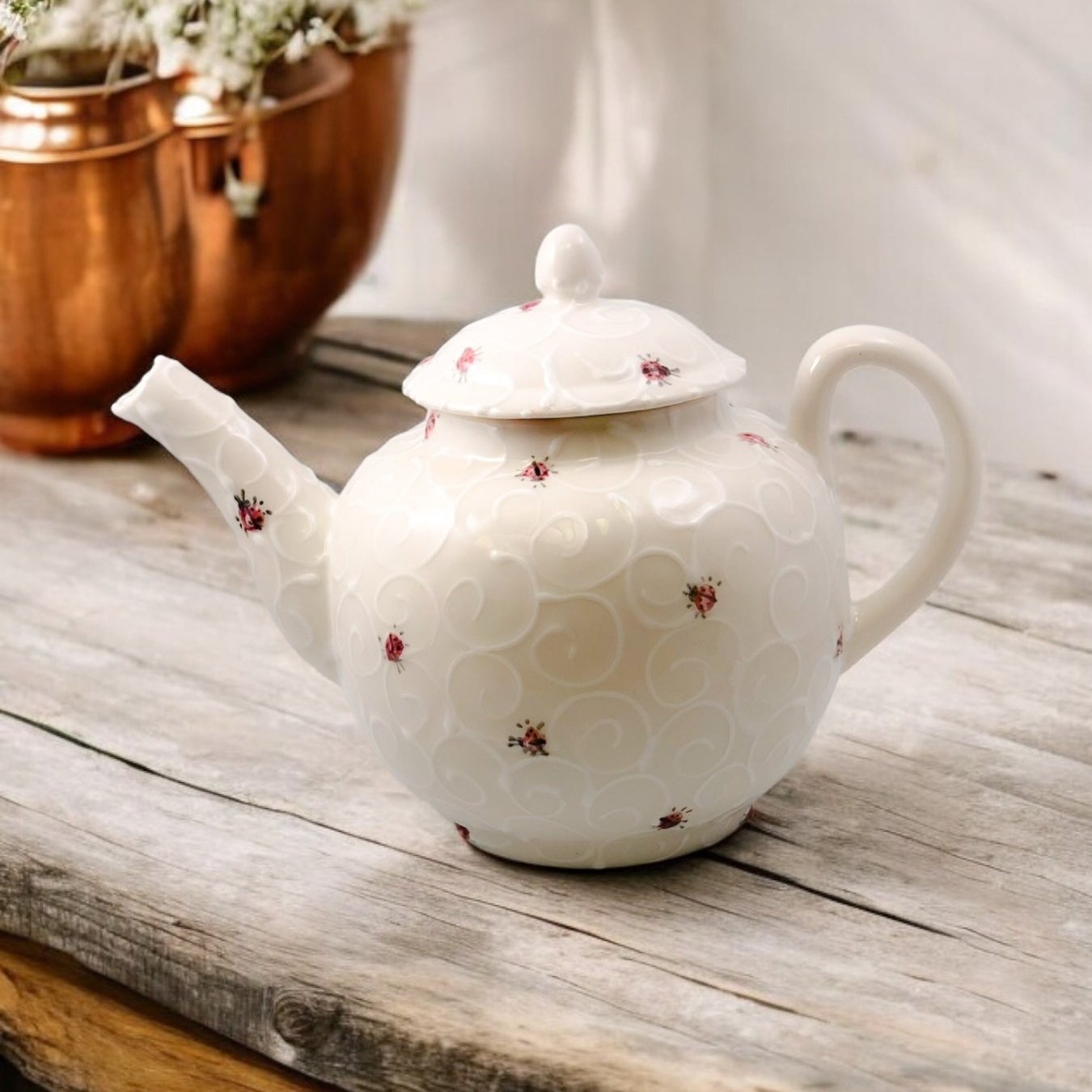 A white ceramic teapot with hand-painted ladybug designs, displayed on a wooden surface with a bouquet of white flowers beside it.