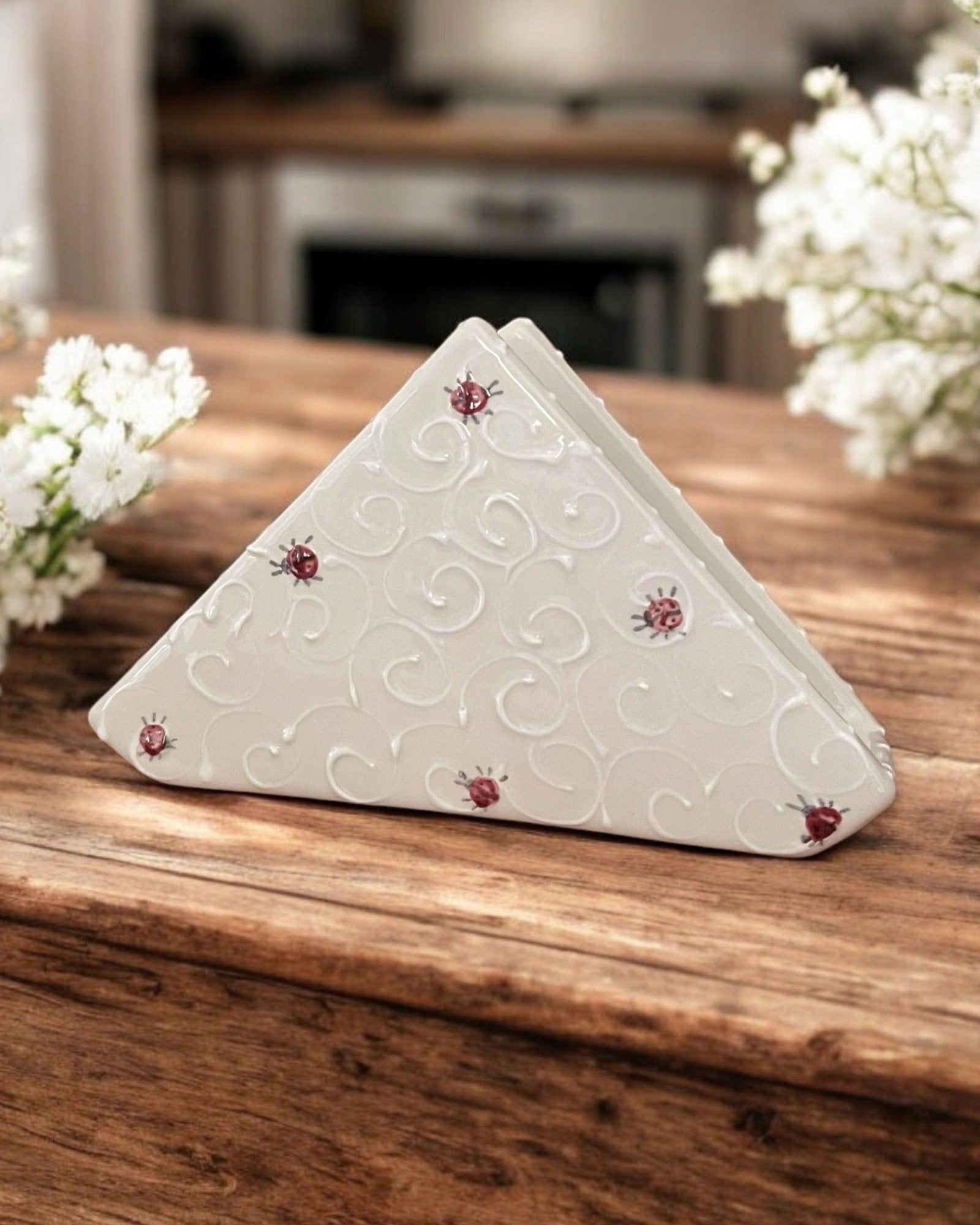 A triangular white ceramic napkin holder with embossed ladybug designs, displayed on a wooden table in front of copper planters filled with purple, blue, and lavender hydrangeas.