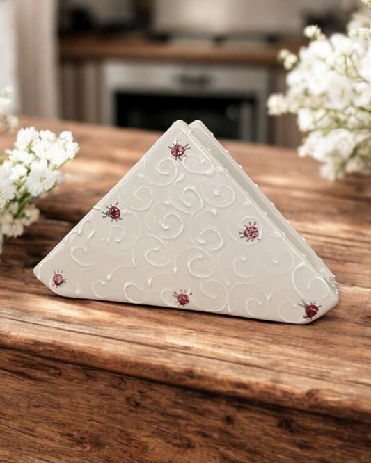 A triangular white ceramic napkin holder with embossed ladybug designs, displayed on a wooden table in front of copper planters filled with purple, blue, and lavender hydrangeas.